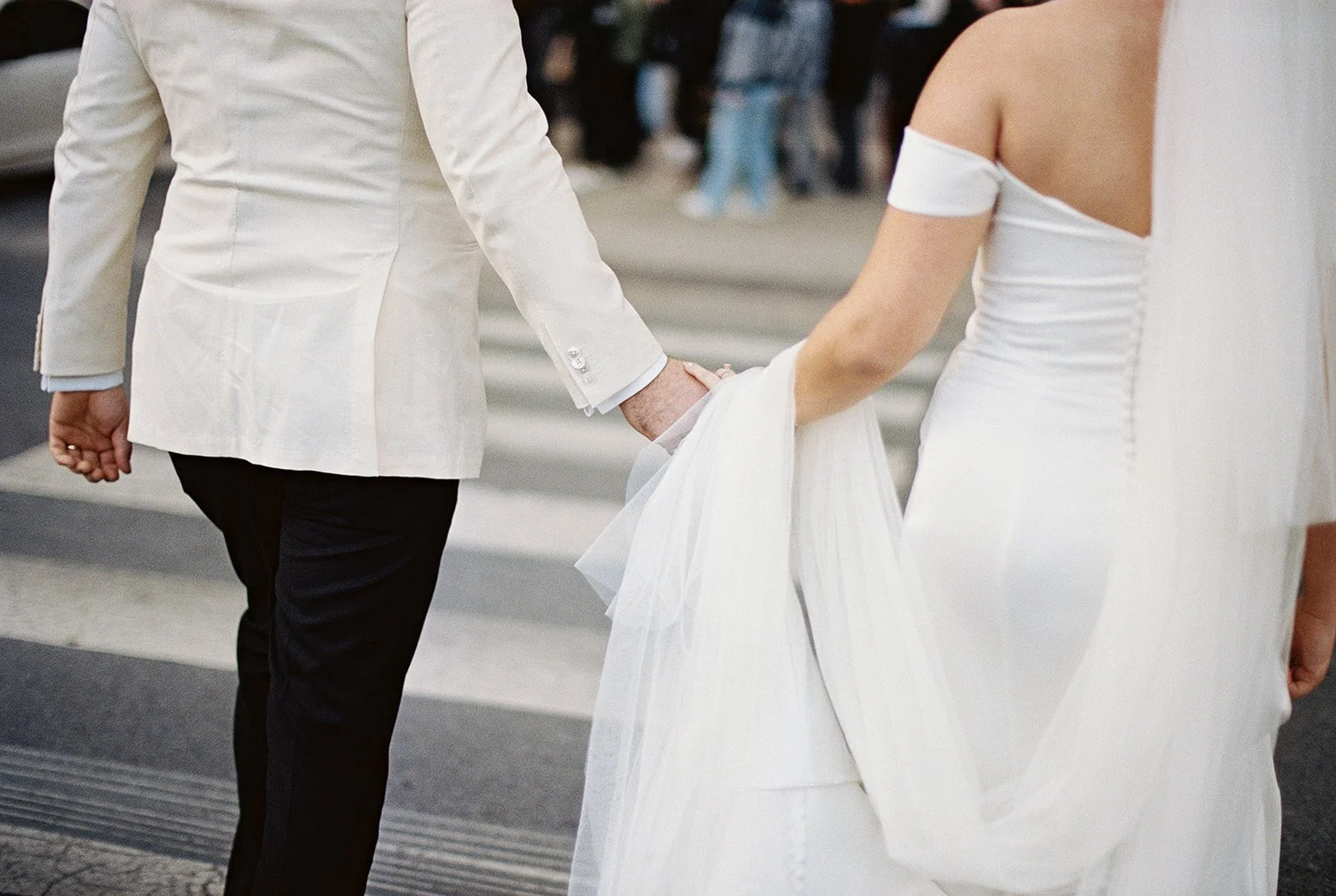 The bride and groom crossing a downtown Chicago street, shot from behind — his white jacket, her cathedral veil and train gathered gently in his hand, the city alive and moving around them.