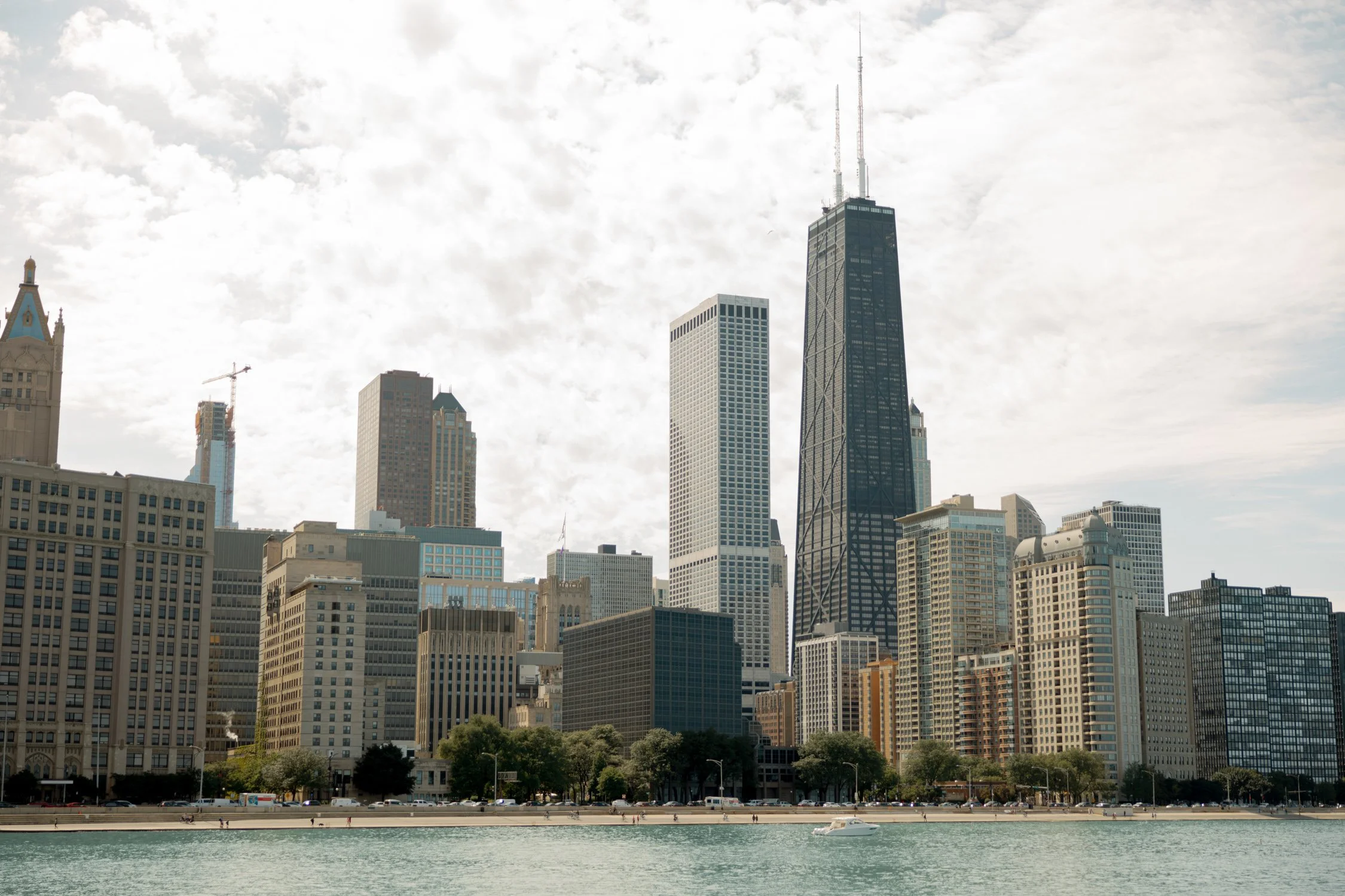 A sweeping view of the Chicago skyline from the Olive Park lakefront, with the iconic John Hancock Tower rising above the calm blue waters of Lake Michigan.
