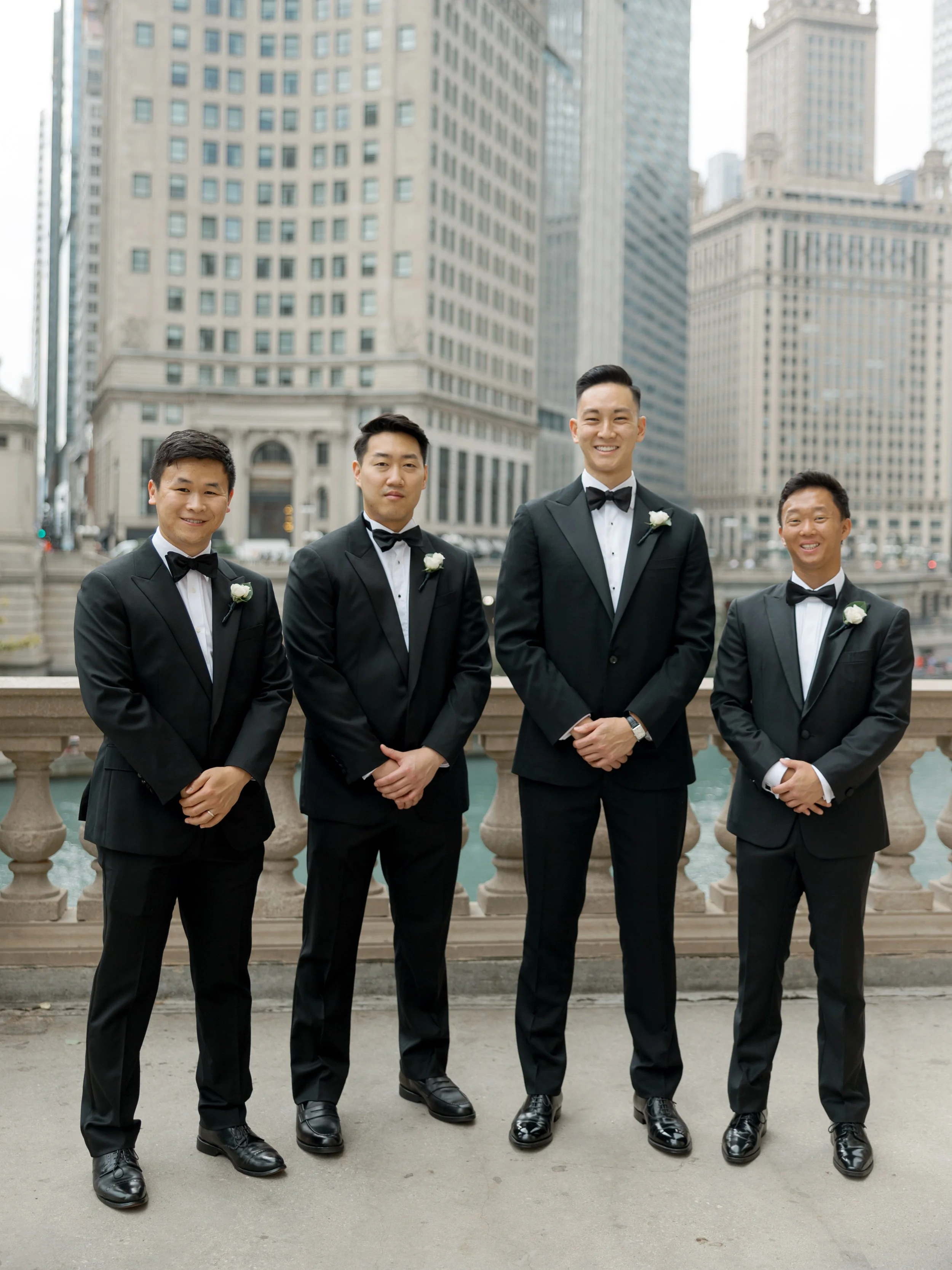 Film portrait of four groomsmen in tuxedos posed on a bridge with the Wrigley Building and Chicago skyline behind them