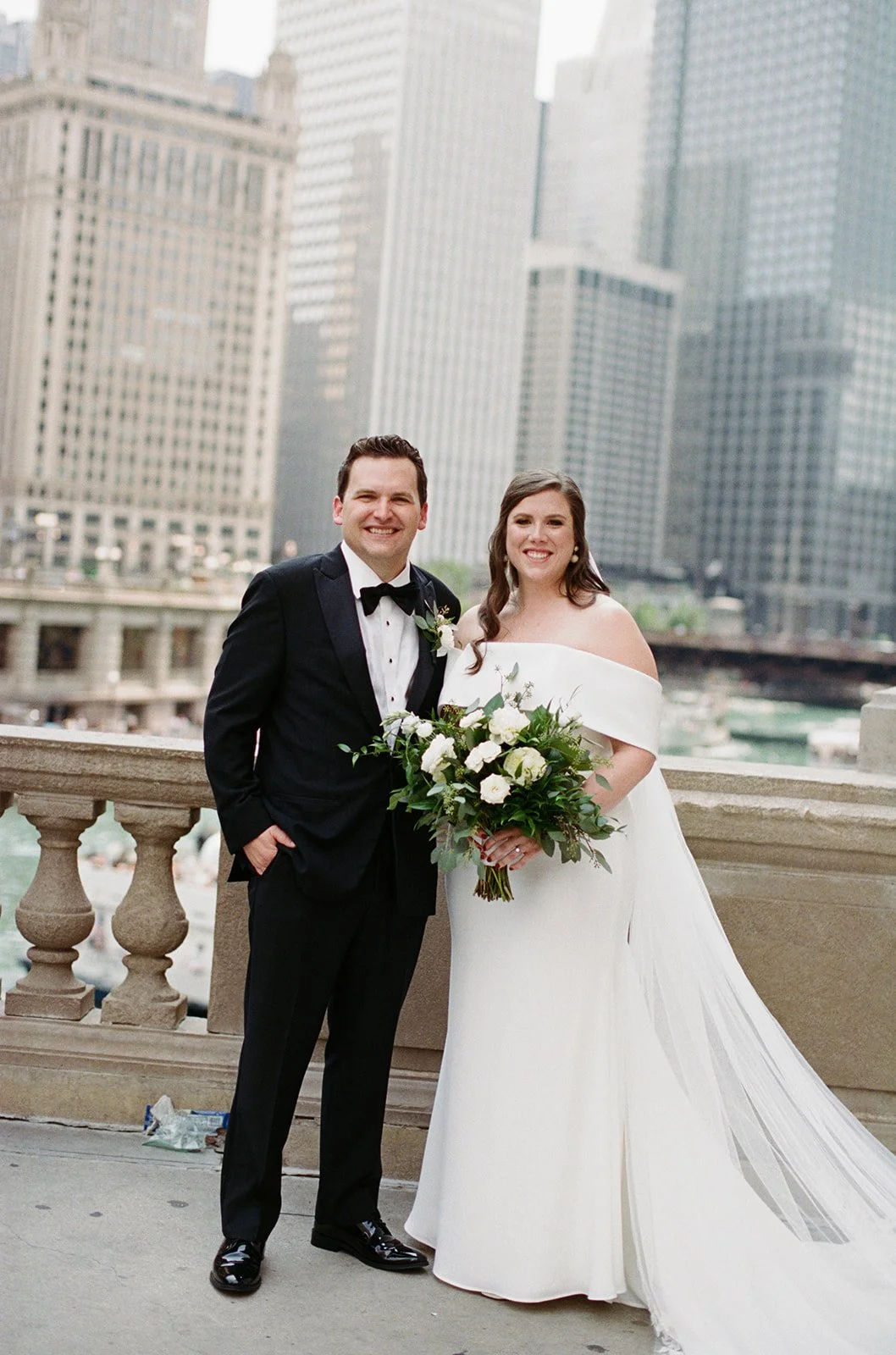 Bride and groom in black tie on the Chicago Riverwalk, city skyline including Wrigley Building and Tribune Tower behind them.