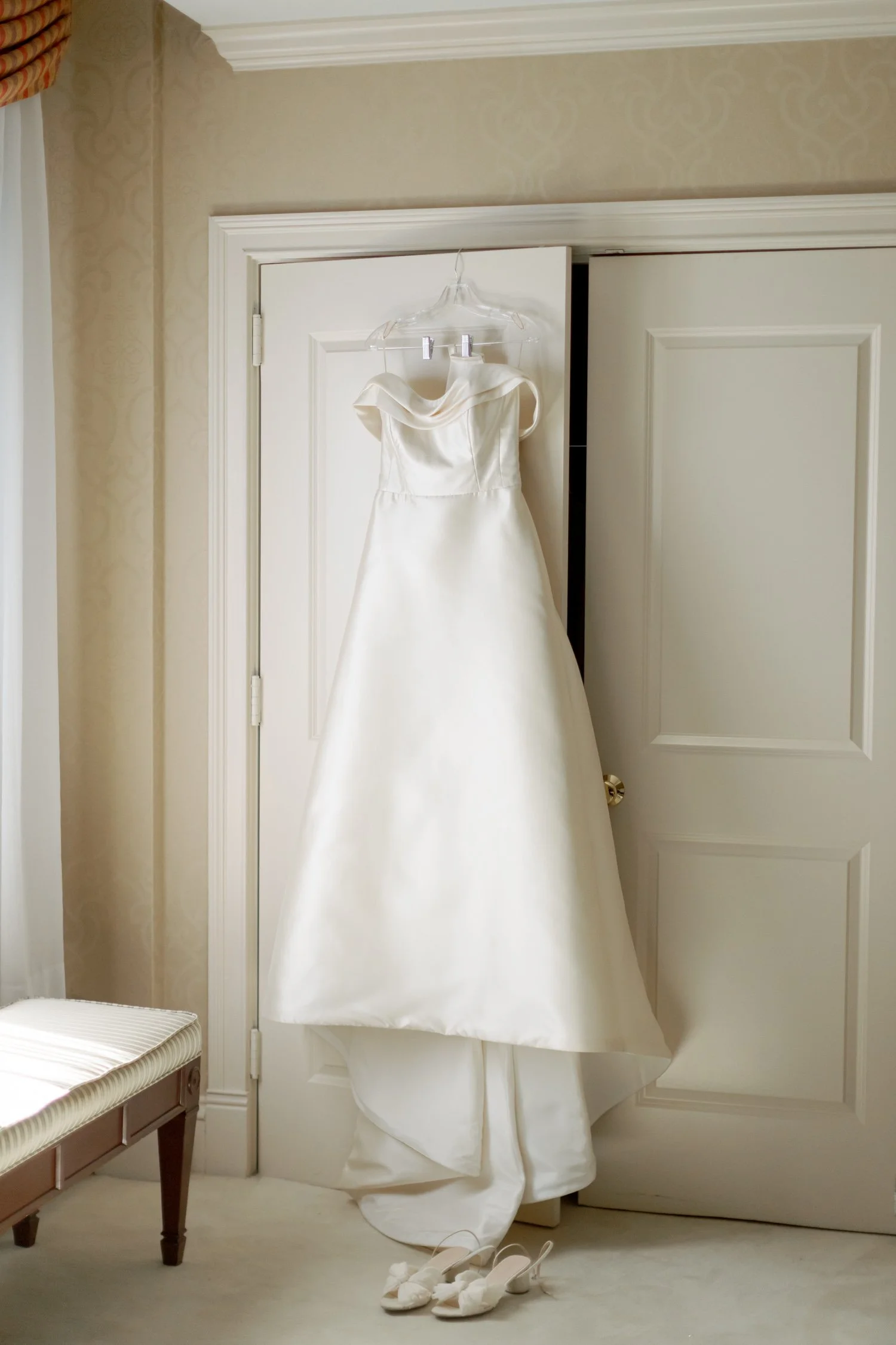 The bride’s sculptural off-shoulder silk gown hangs from a white hotel closet door, her bridal shoes resting just below on the carpet — quiet and still.