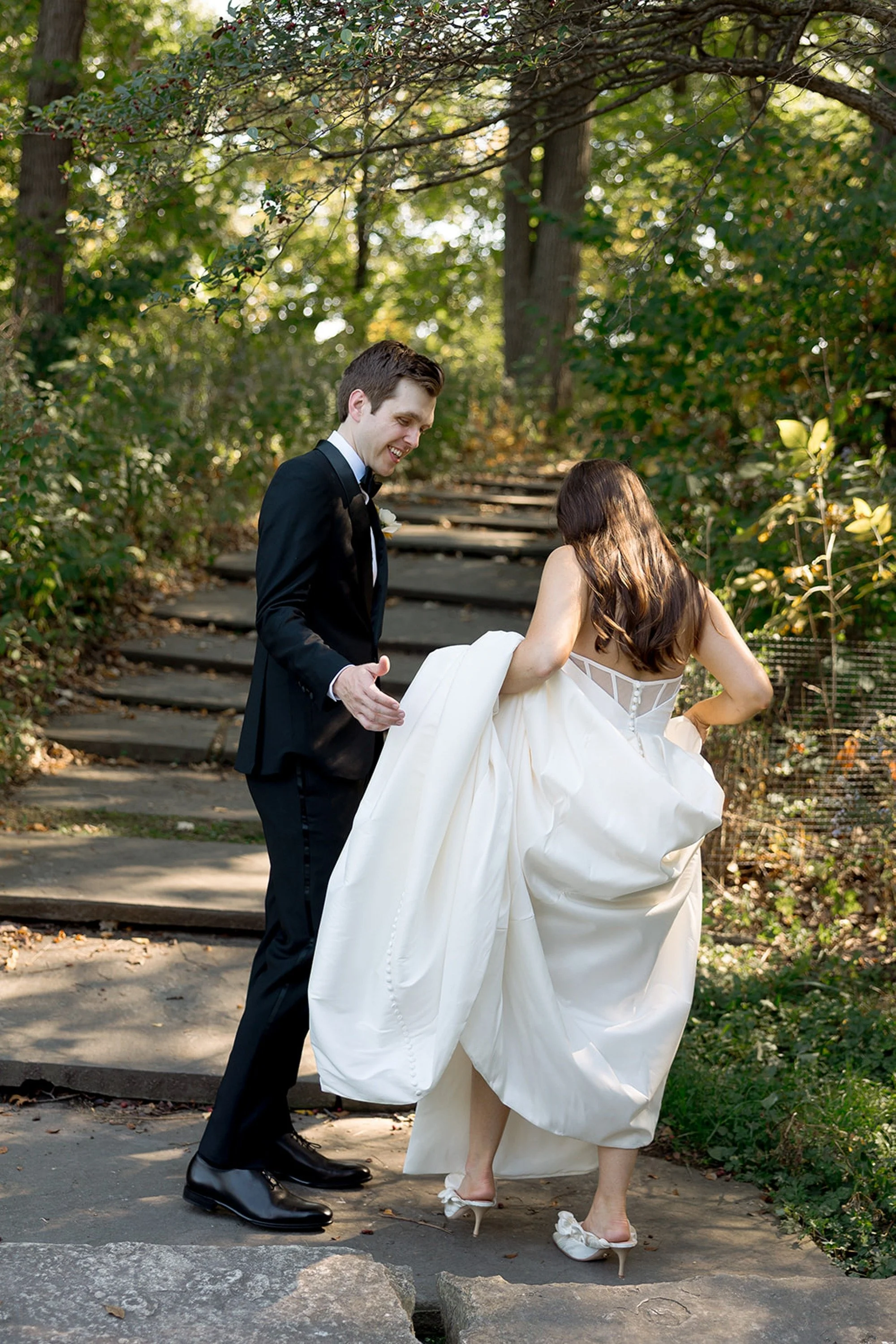 The groom steadies his bride as she lifts her voluminous ballgown on stone garden steps — a genuine, playful moment on the wooded grounds of Columbus Park Refectory. Chicago film wedding photographer.