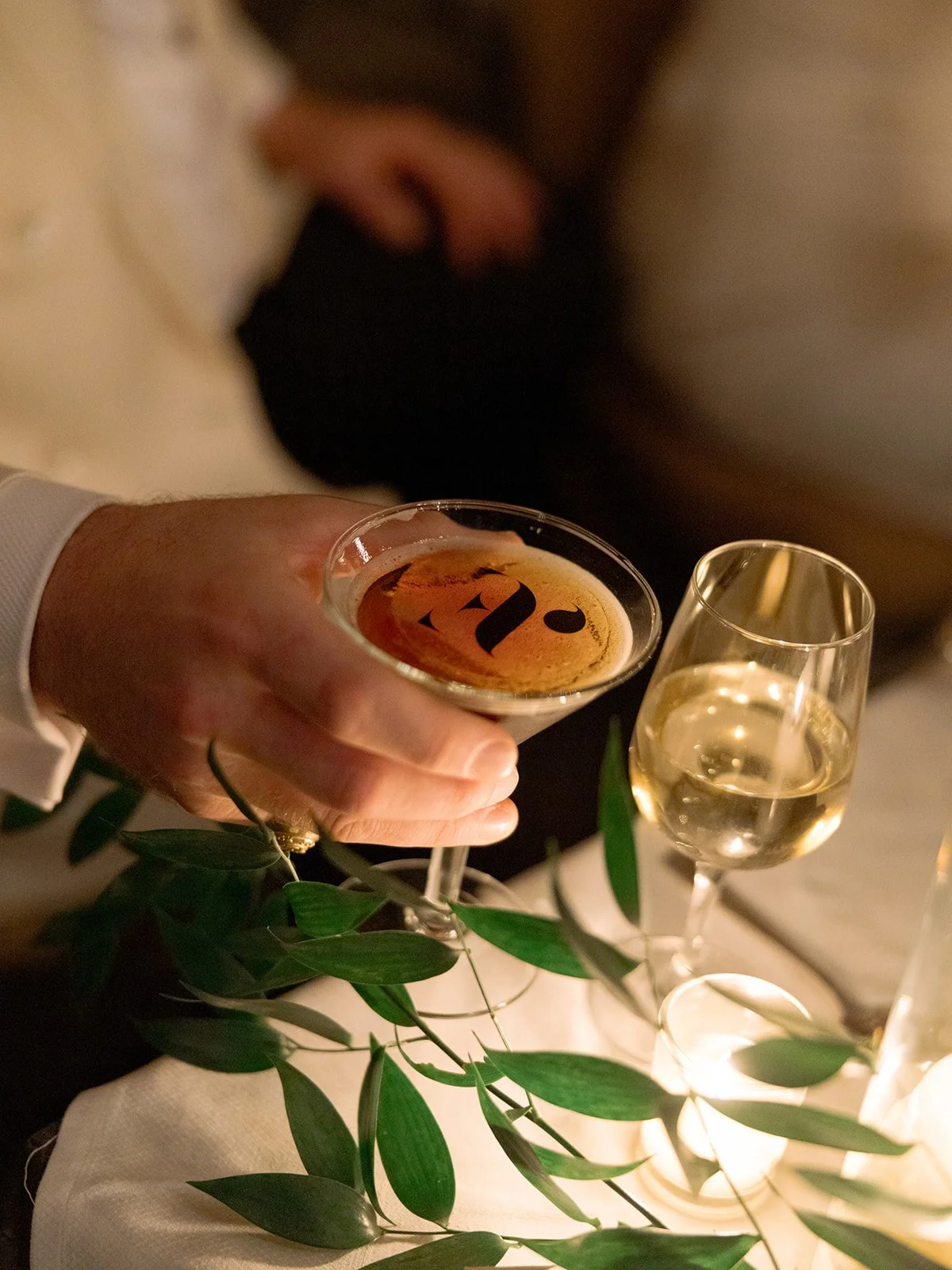 The groom holds a monogrammed espresso martini at the sweetheart table, the lush greenery-and-floral installation above the fireplace softly framing him behind.