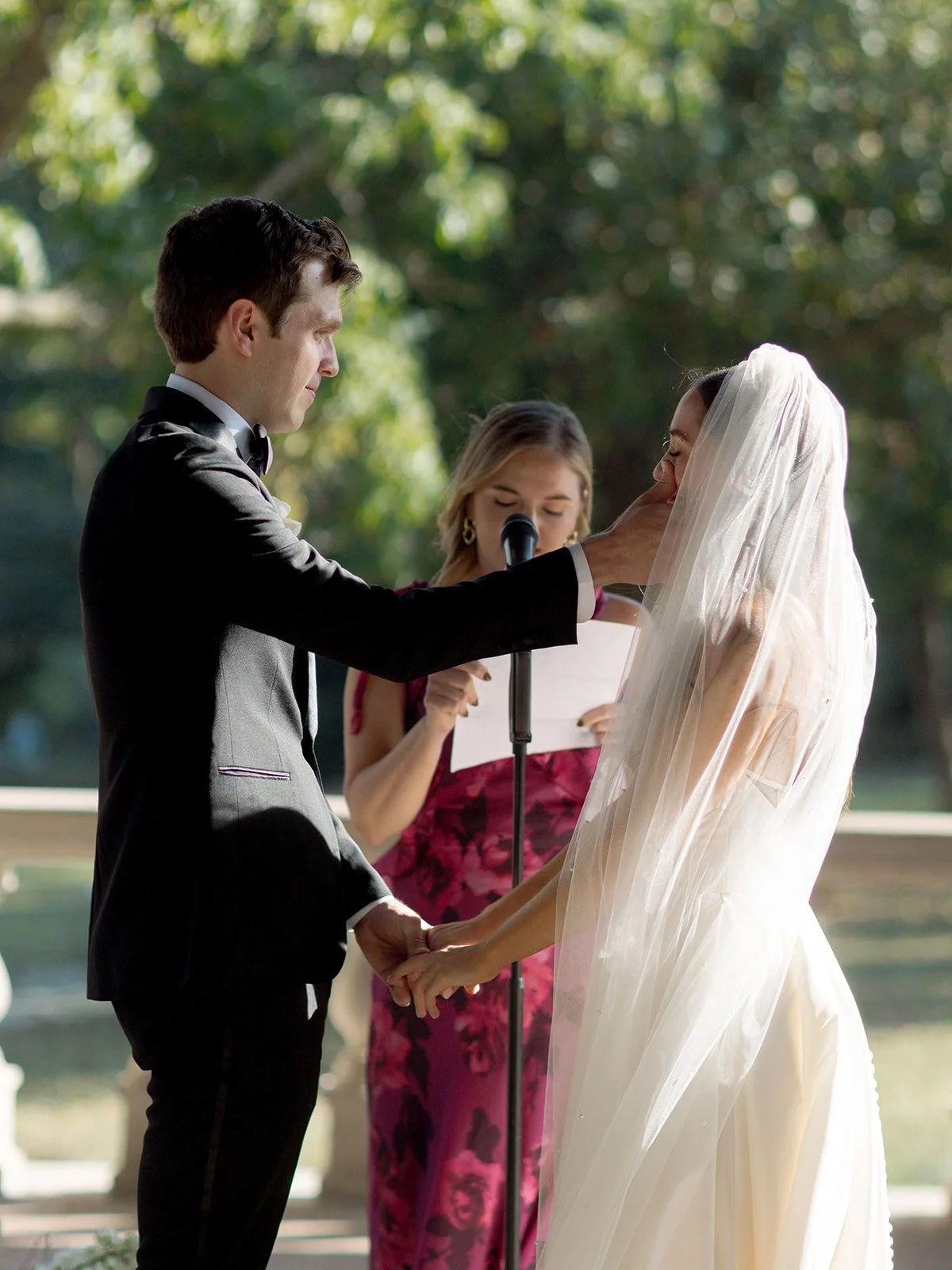 The groom reads his vows from paper, one hand gently lifting his bride's veil in a luminous outdoor ceremony moment. The officiant stands behind them in a garden of green — Columbus Park Refectory, Chicago film wedding photographer.