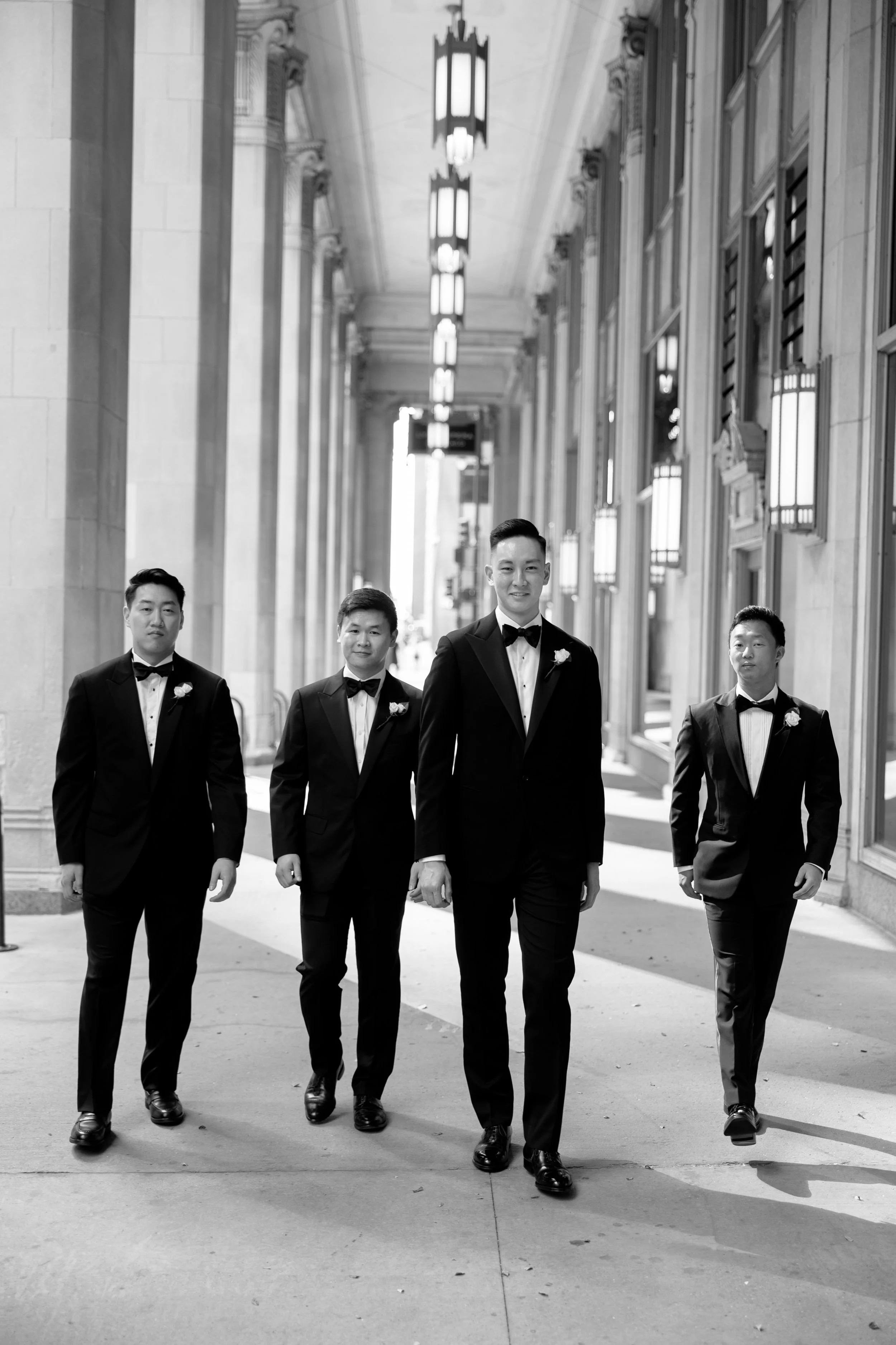 Black and white film portrait of four groomsmen in tuxedos walking down the Lyric Opera colonnade in Chicago
