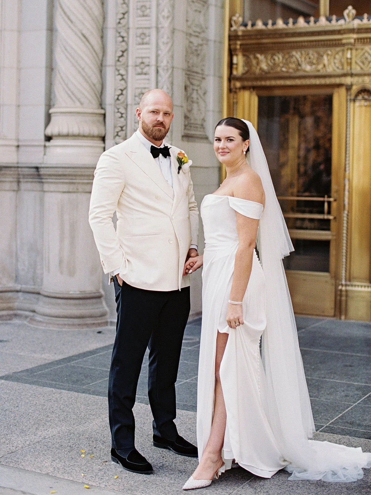 Bride and groom holding hands outside the ornate gold entrance of a historic Chicago building — relaxed, confident, and completely at ease in front of the camera. The bride's gown slit and low heels give the whole thing a downtown-cool feel