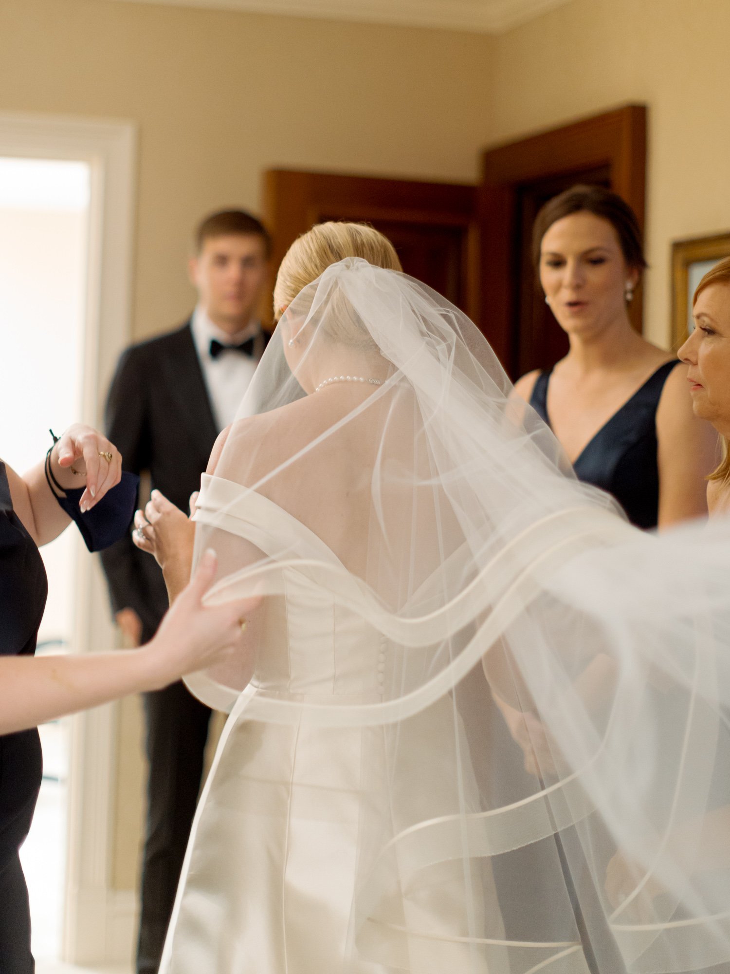 The bride’s cathedral veil billows dramatically mid-air as attendants carefully arrange it around her shoulders just moments before the ceremony begins.