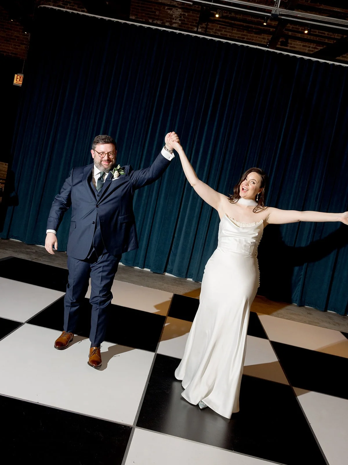 Bride in a silk slip gown and groom in a navy suit raising hands on a black and white checkered dance floor during their grand entrance at Wildman BT in Chicago, photographed by Louie Abellera.
