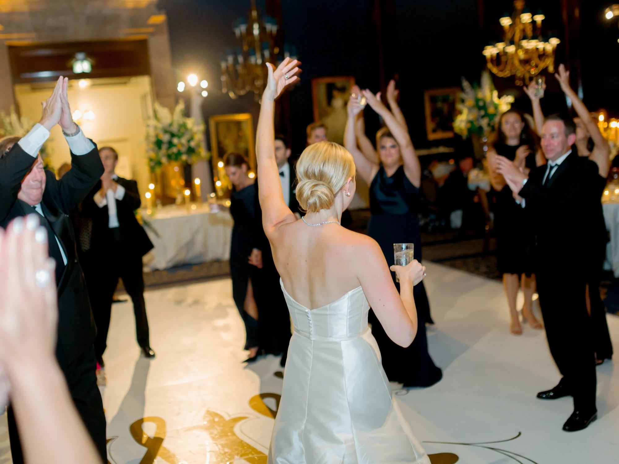 The bride raises both arms toward the crowd on the reception dance floor, the room responding around her — every great party has a moment like this.
