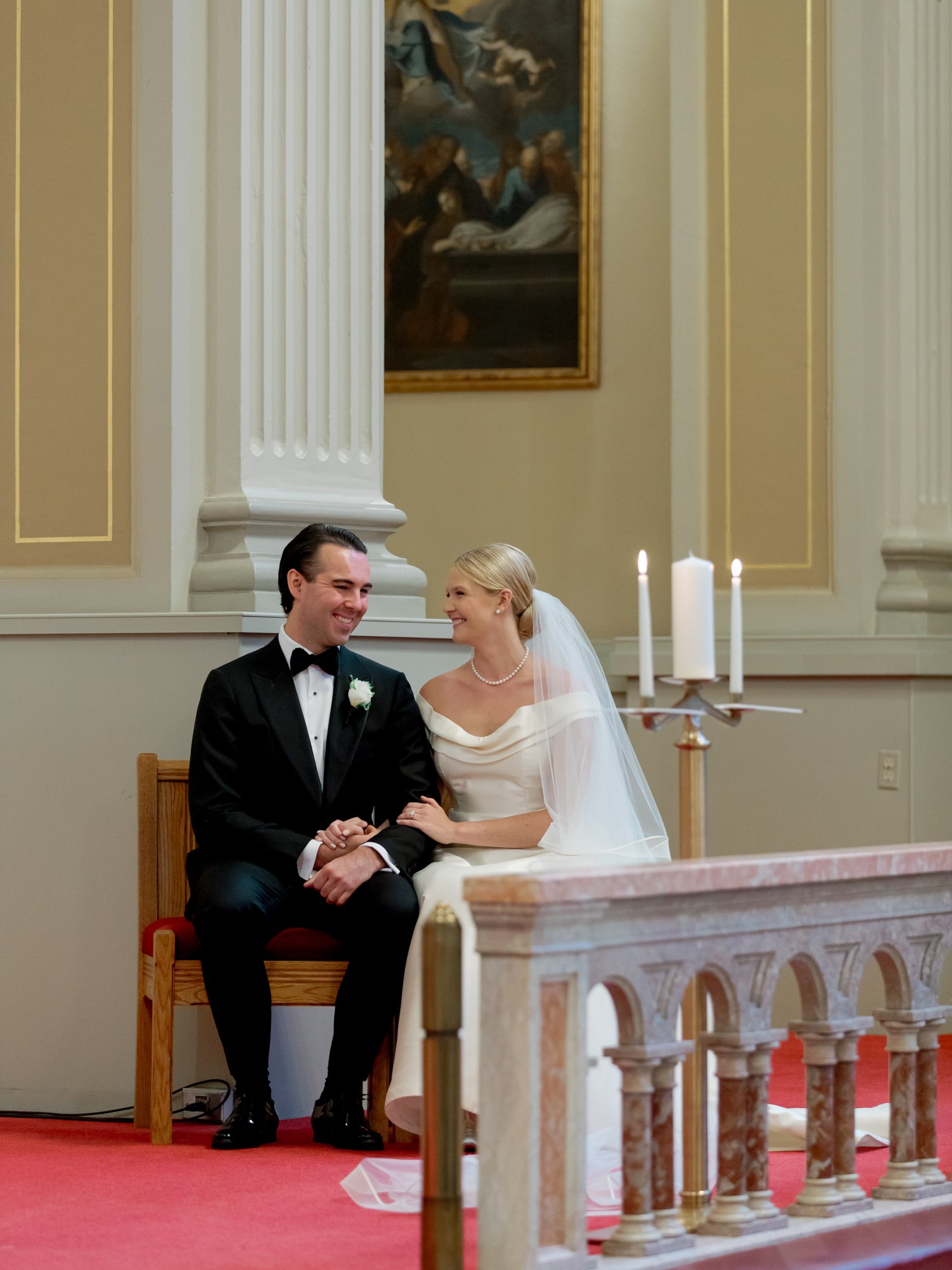 Bride and groom share a quiet, smiling glance seated at the ceremony altar in a classical Chicago chapel, pillar candles and columns rising behind them.
