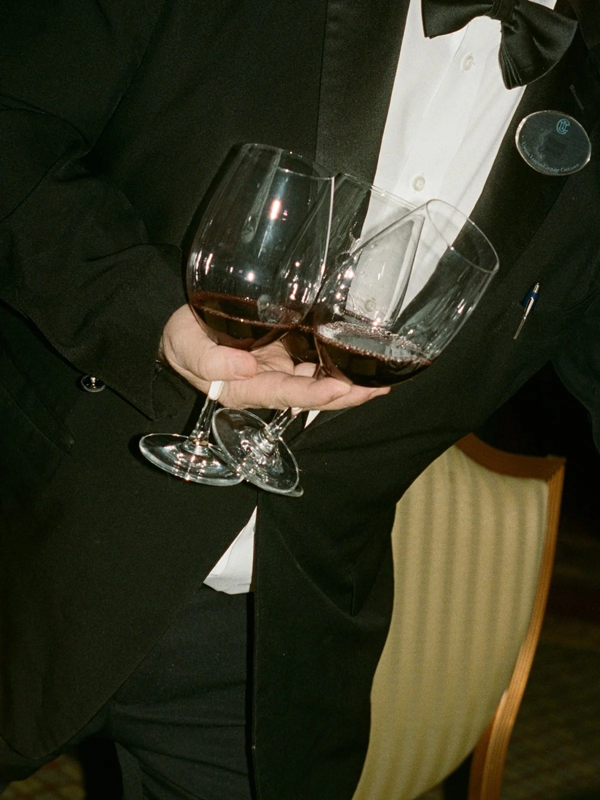 Film close-up of a tuxedoed waiter carrying two glasses of red wine at a wedding reception at the Union League Club of Chicago, photographed by Louie Abellera.