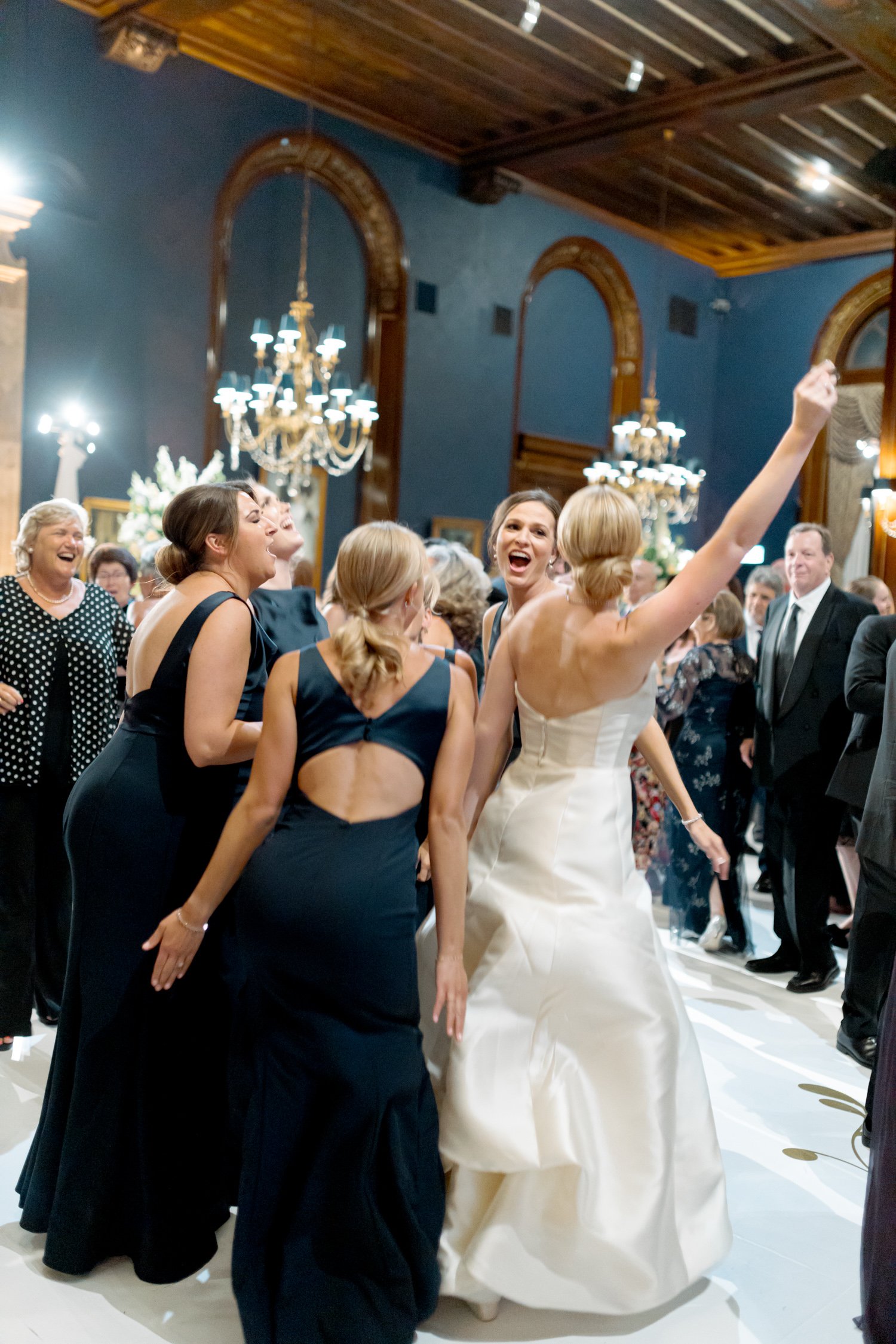 Bride raises her arm and laughs freely mid-dance in her strapless silk gown, surrounded by bridesmaids in black beneath the Union League Club chandeliers.