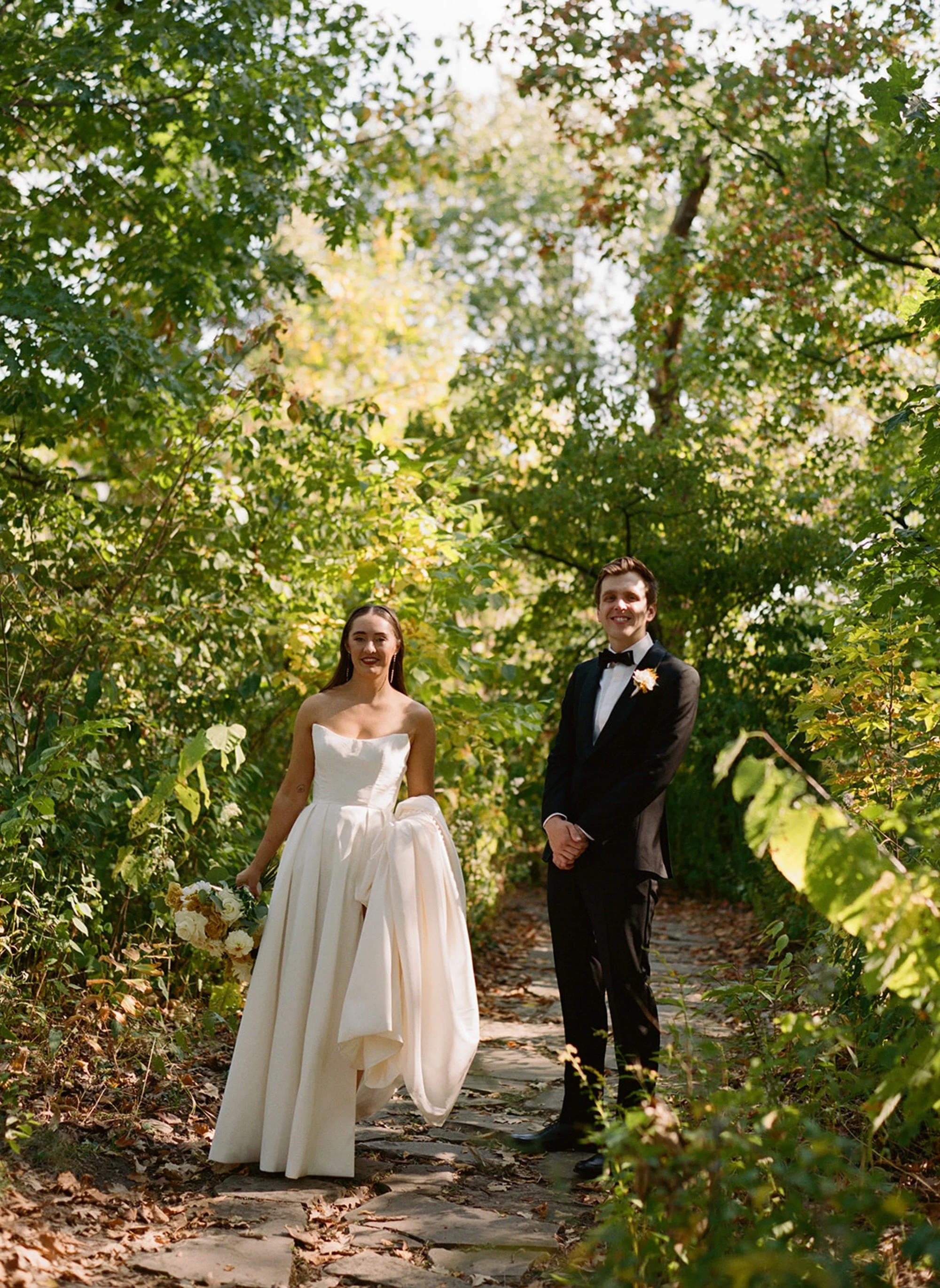 Hand in hand, the couple strolls through a verdant autumn garden tunnel on the grounds of Columbus Park Refectory — the bride's gown catching the breeze, the groom glancing back at her. Chicago film wedding photographer.