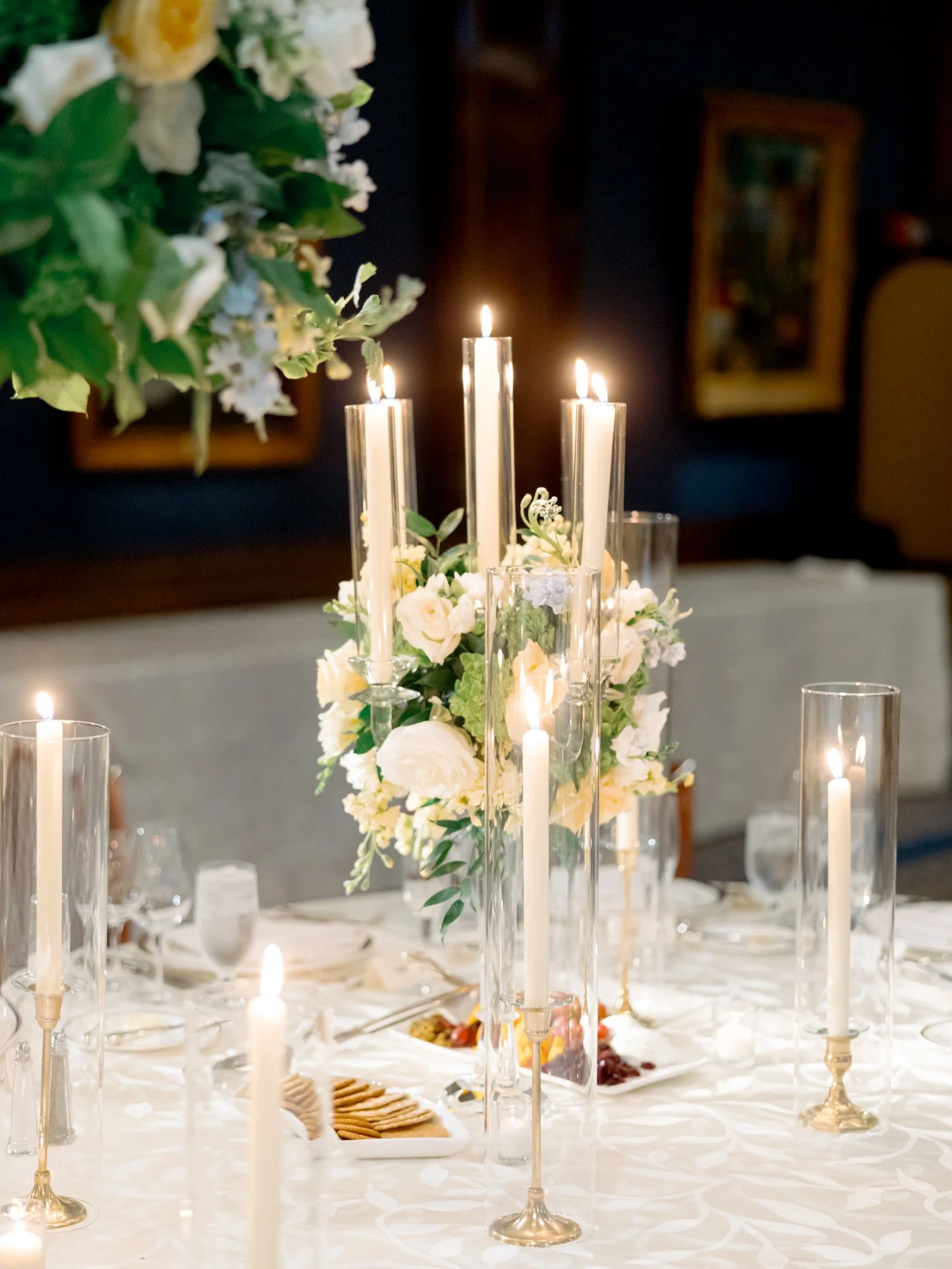 Tall glass taper candles cluster around a white and yellow rose centerpiece on the reception table, the deep navy Union League Club dining room behind.