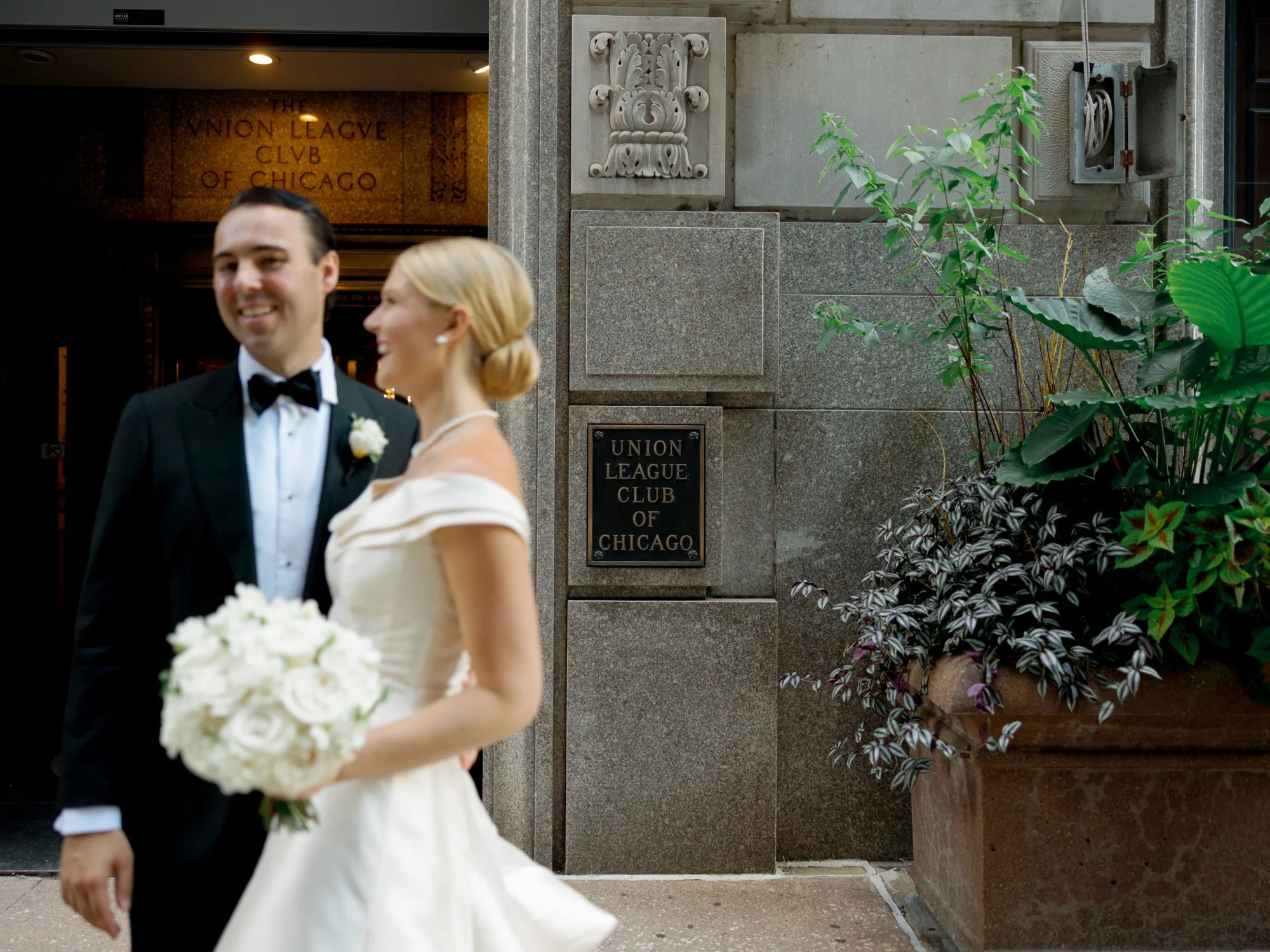 Bride and groom walk and laugh together just outside the Union League Club entrance, the iconic bronze plaque visible on the stone wall behind them.