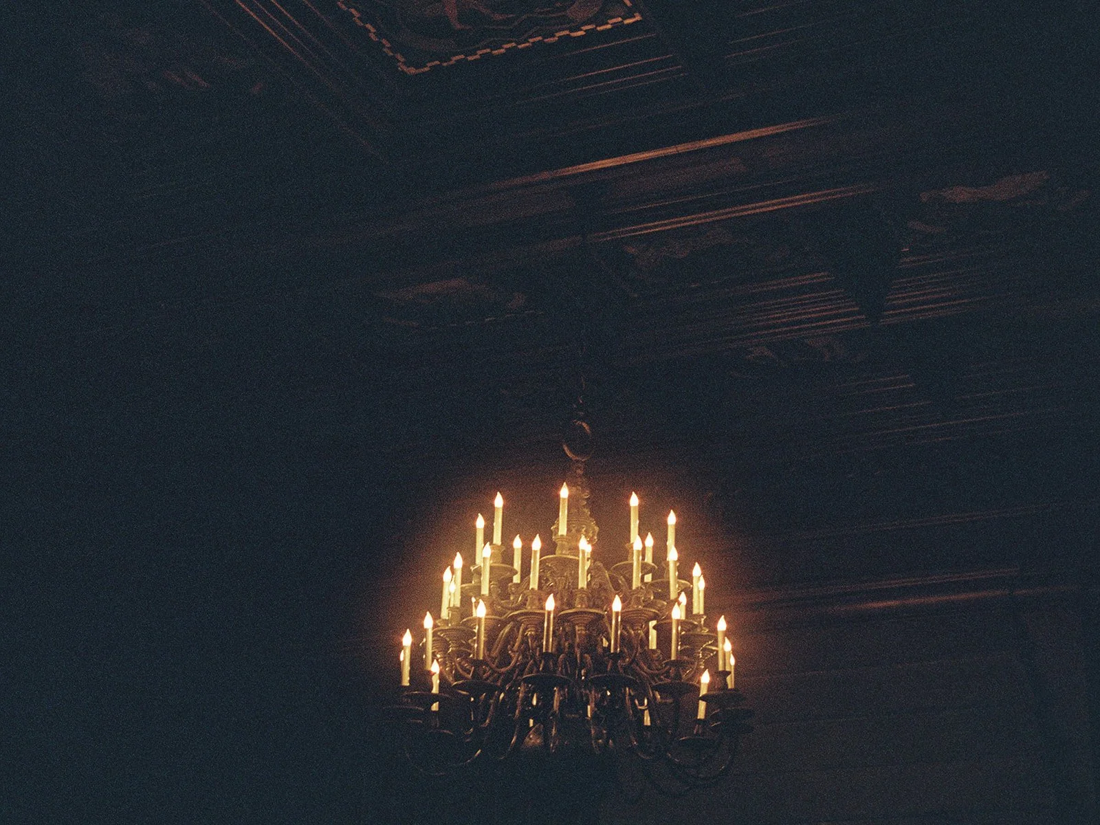 A moody, cinematic shot of the University Club Chicago's candelabra-style chandelier glowing against the dark room, ornate carved ceiling barely visible above. Pure atmosphere — the kind of frame that sets the tone for the whole gallery.
