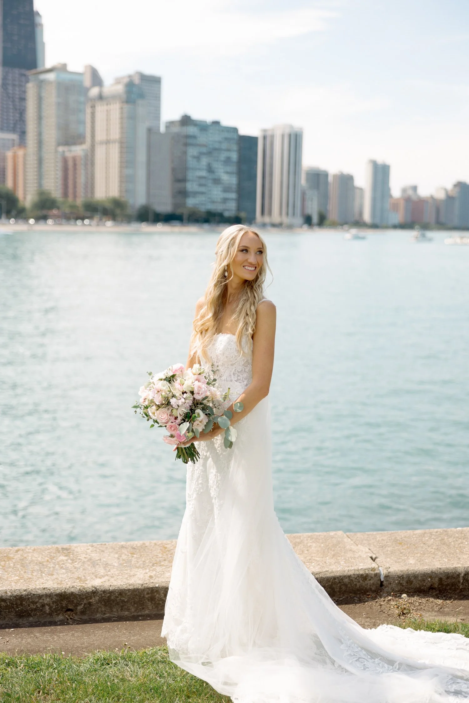 The bride stands radiant at Olive Park, her lace gown and pink bouquet glowing against the turquoise lake and iconic Chicago skyline behind her.