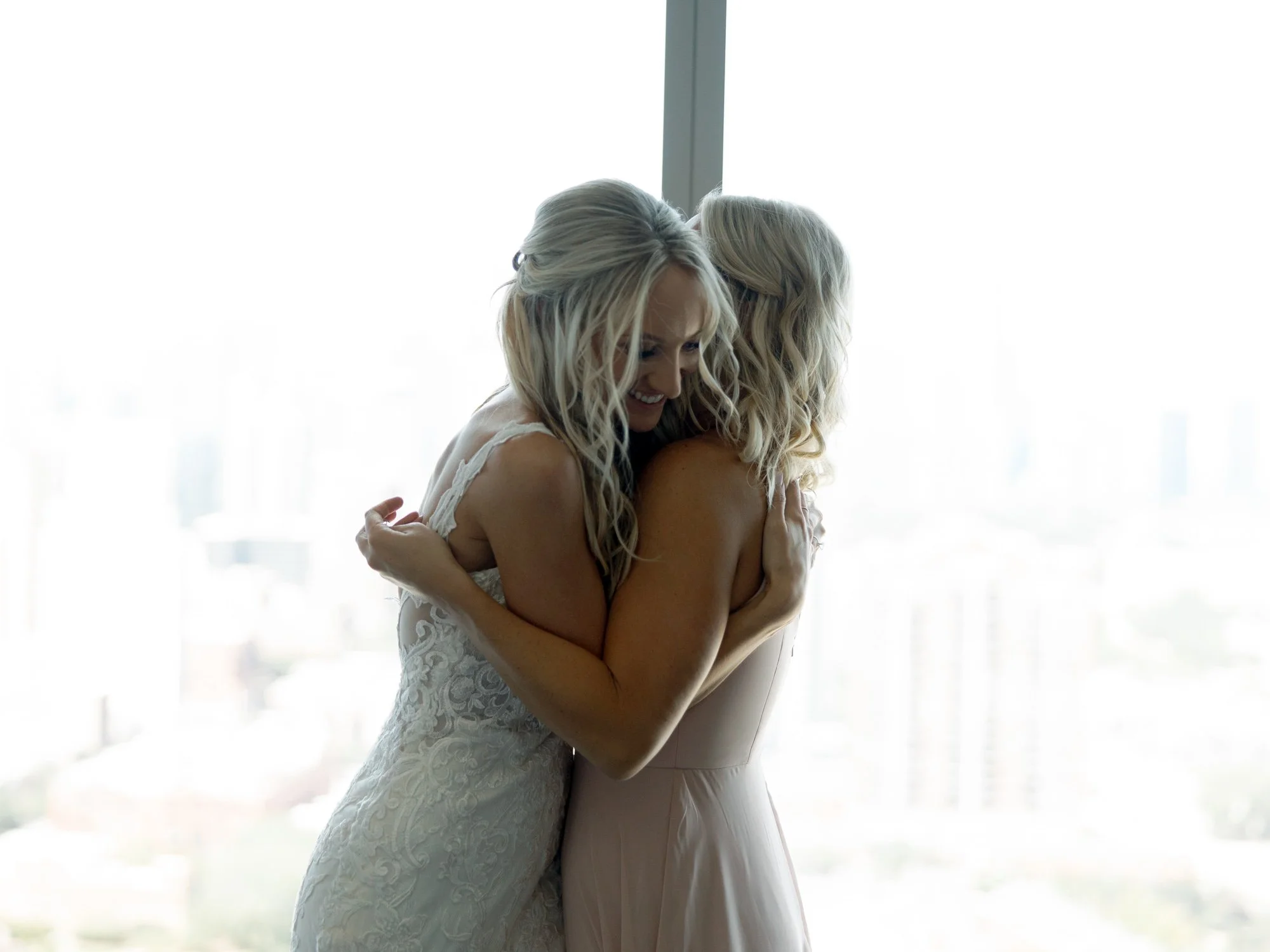 The bride and bridesmaid share a heartfelt embrace silhouetted by floor-to-ceiling windows, the soft Chicago skyline glowing gently in the background.