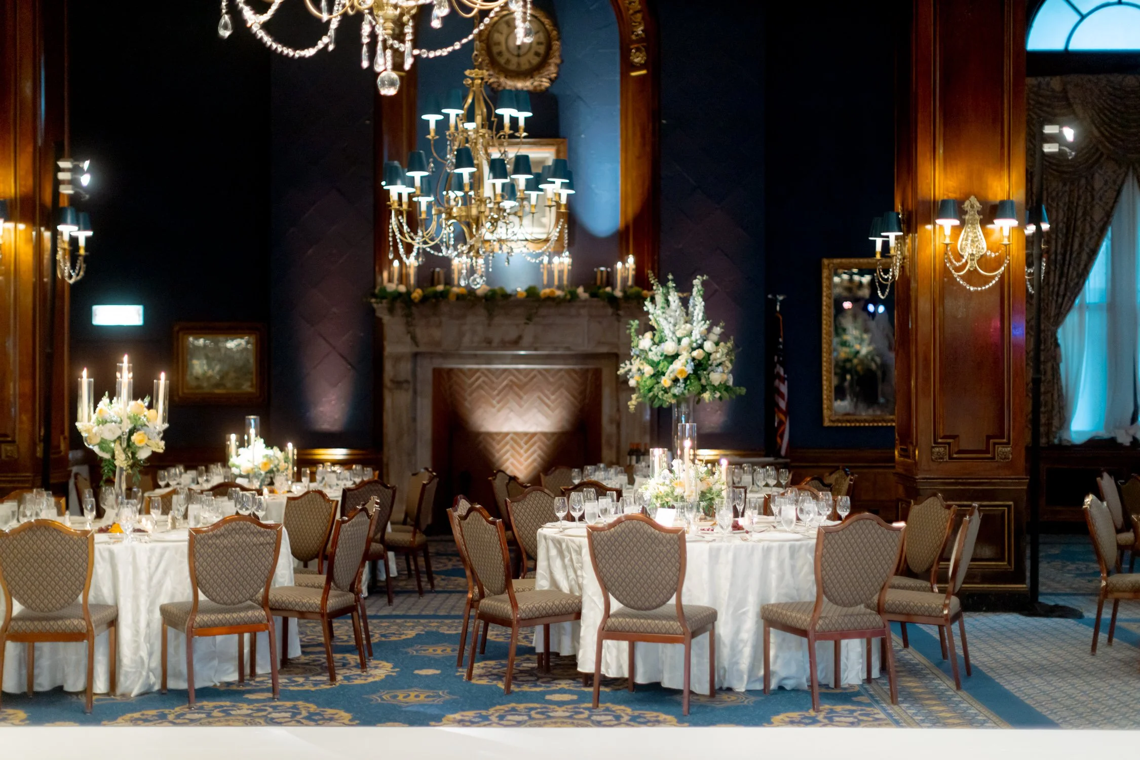 Wide shot of the Union League Club reception room fully set — round tables, ornate chandeliers overhead, and a carved marble fireplace at the far wall.
