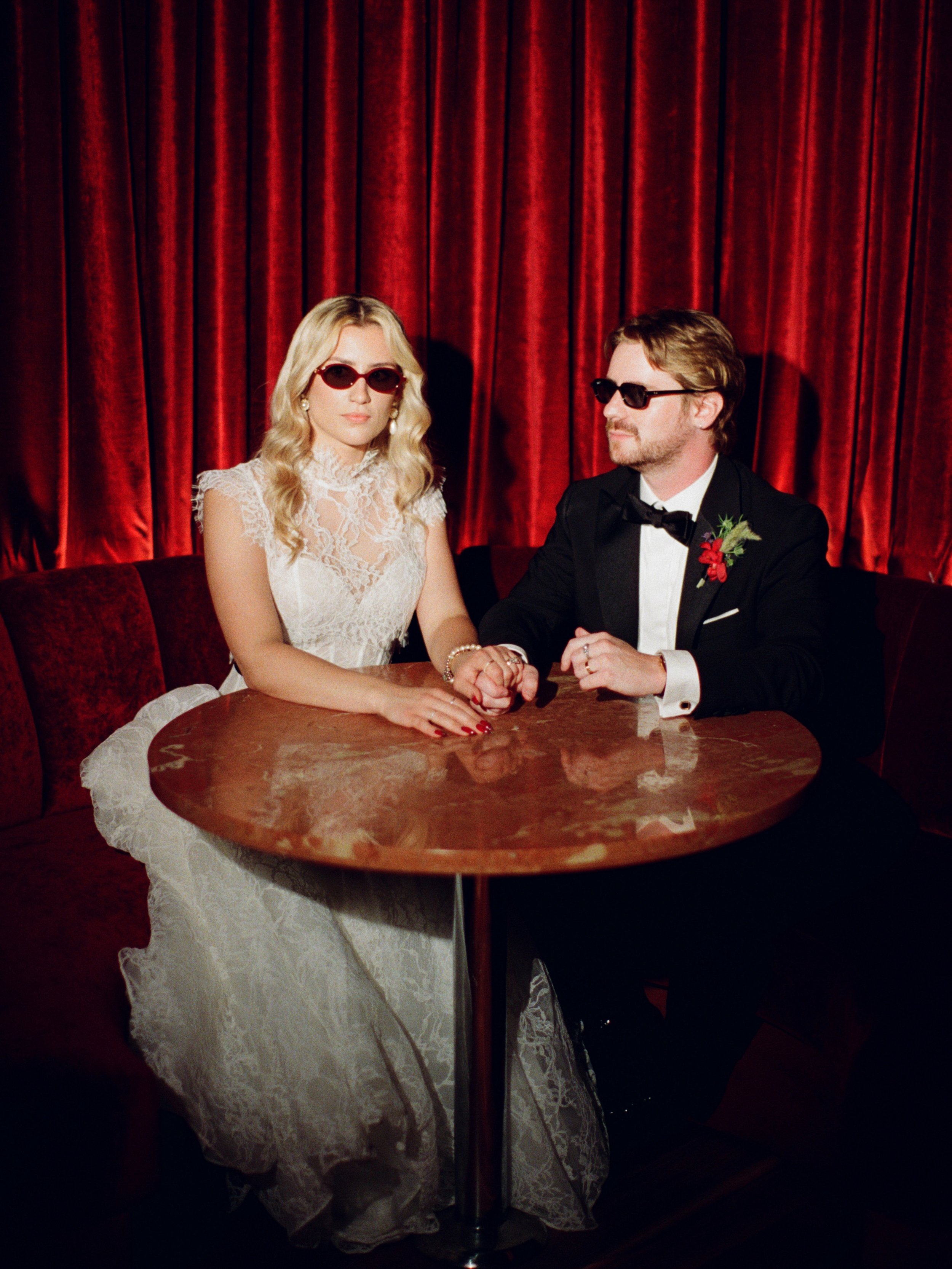 Bride and groom pose in sunglasses at their round table inside The Wellsley's red velvet lounge