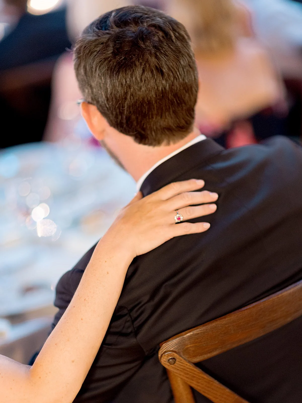 Close-up of the bride's hand resting on the groom's shoulder at the reception, wedding rings catching warm light.