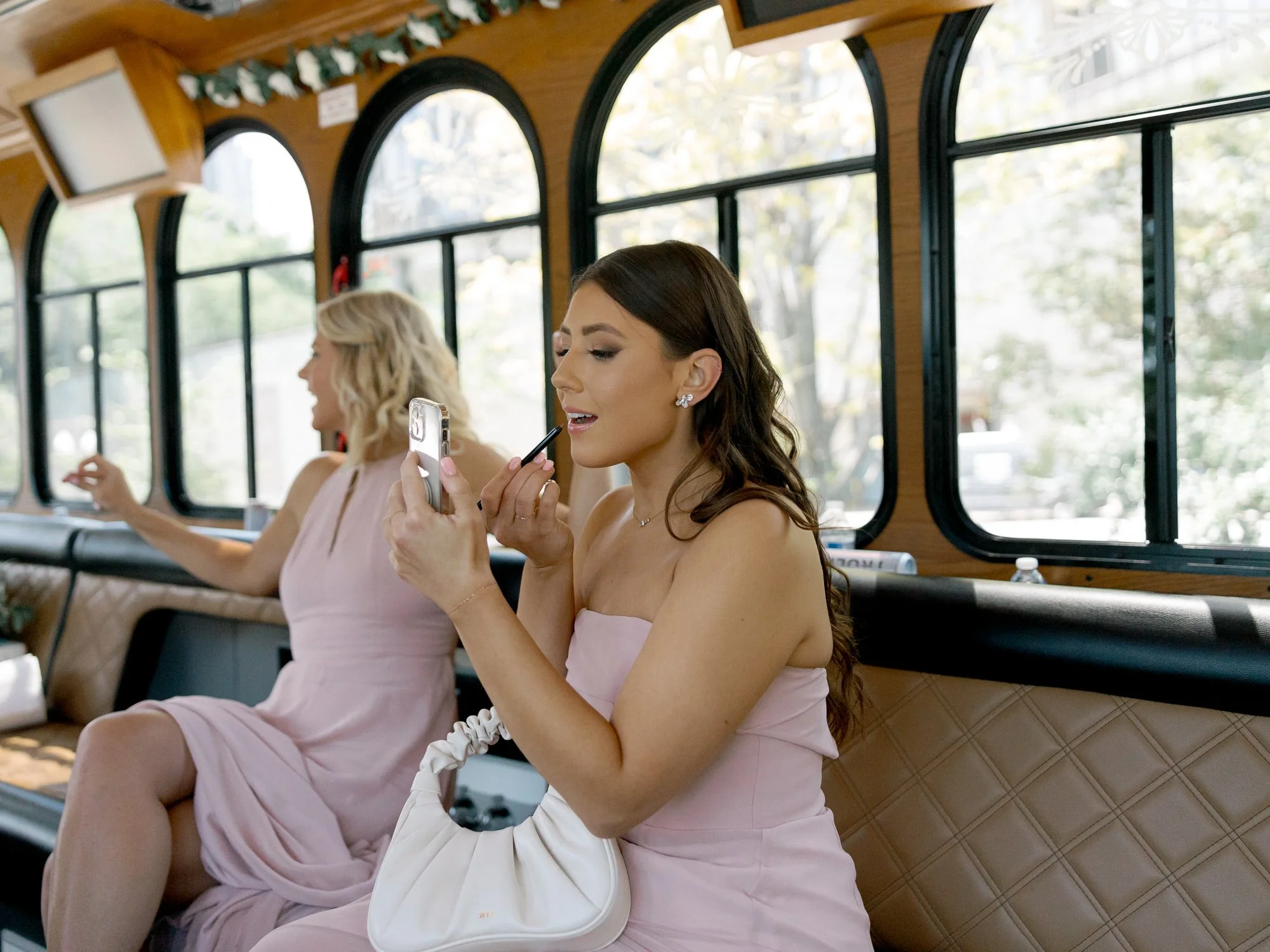 A bridesmaid in blush touches up her lipstick on the wedding trolley, while her companion smiles beside her in the warm afternoon light.