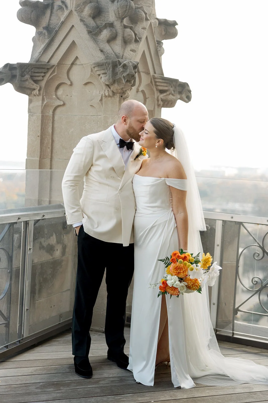 The groom kisses his bride's temple on the University Club Chicago's Gothic rooftop terrace, a stone spire rising behind them and the city stretching into the horizon. Tender and sweeping all at once — by a Chicago film wedding photographer.