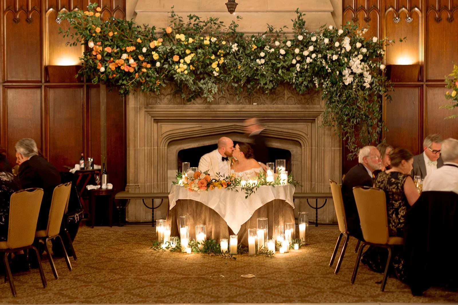 The University Club Chicago ballroom in full reception mode — the couple kissing at their sweetheart table in the center, guests gathered at candlelit round tables, the floral arch blooming above the fireplace behind them.