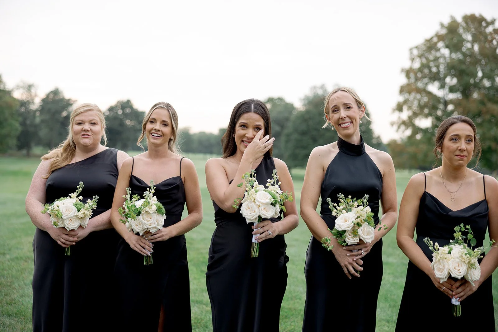 Candid portrait of five bridesmaids in black satin gowns holding white rose bouquets, one wiping away tears, on the grounds of the Glen View Club, photographed by Chicago wedding photographer Louie Abellera.