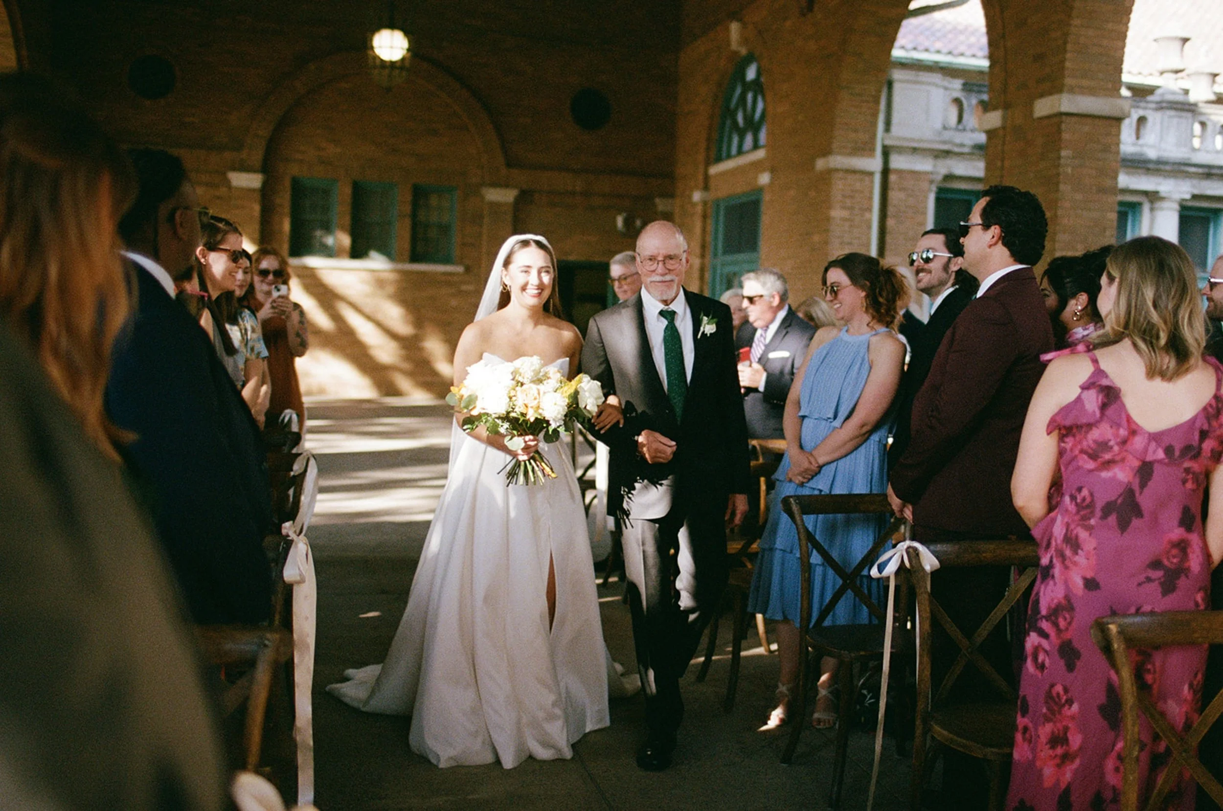 The processional — the bride and her father walking the aisle together as guests rise on either side, sunlight flooding the pavilion of Columbus Park Refectory. Shot on film by a Chicago film wedding photographer