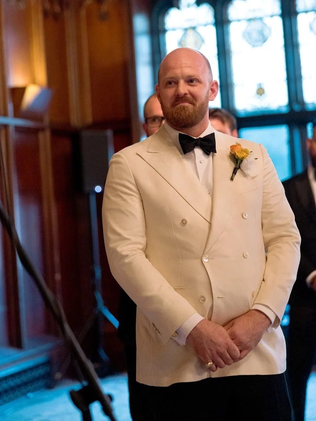 The groom stands at the altar inside the University Club Chicago chapel — hands folded, stained glass windows glowing warmly behind him, the quiet before everything begins.