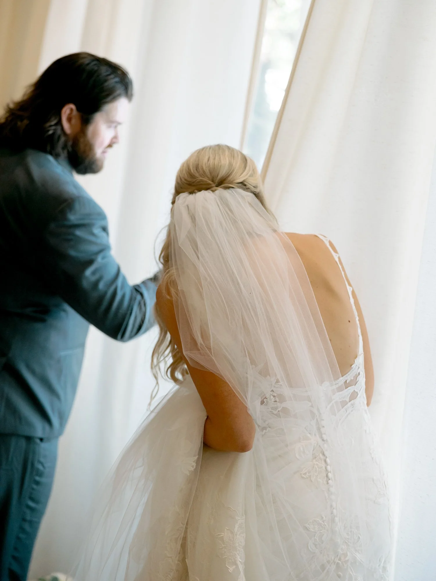 A tender getting-ready moment as someone gently adjusts the bride's cathedral veil by a bright window, her open-back lace gown softly illuminated.