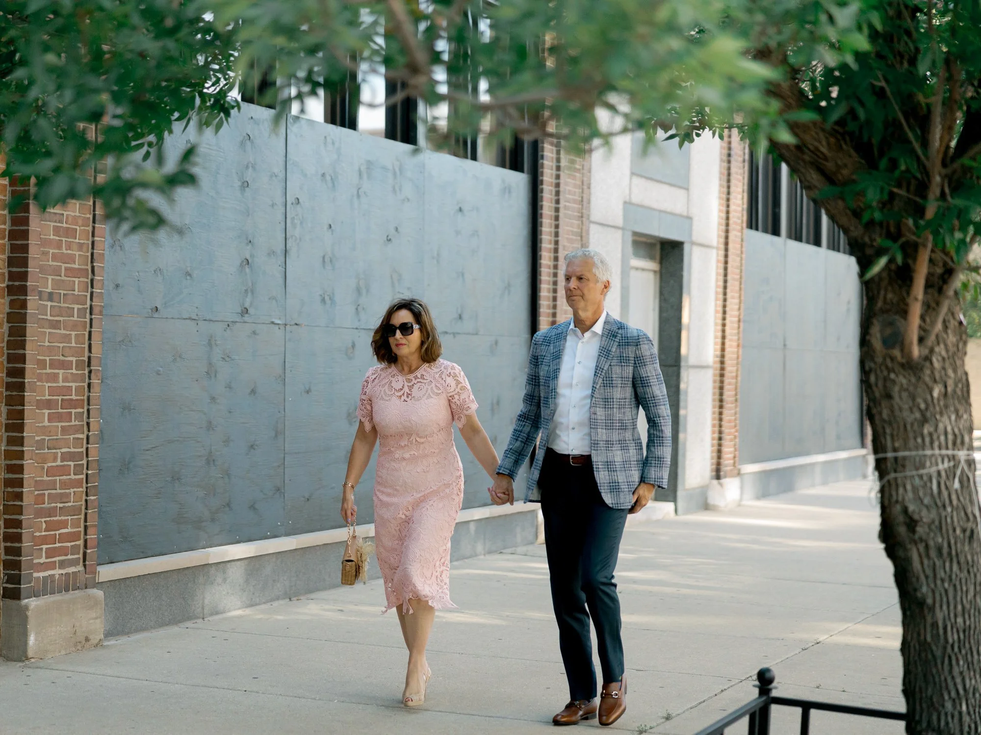 An elegantly dressed couple strolls hand-in-hand on a sun-dappled sidewalk near the Chicago Illuminating Company, her blush lace dress glowing in the light.