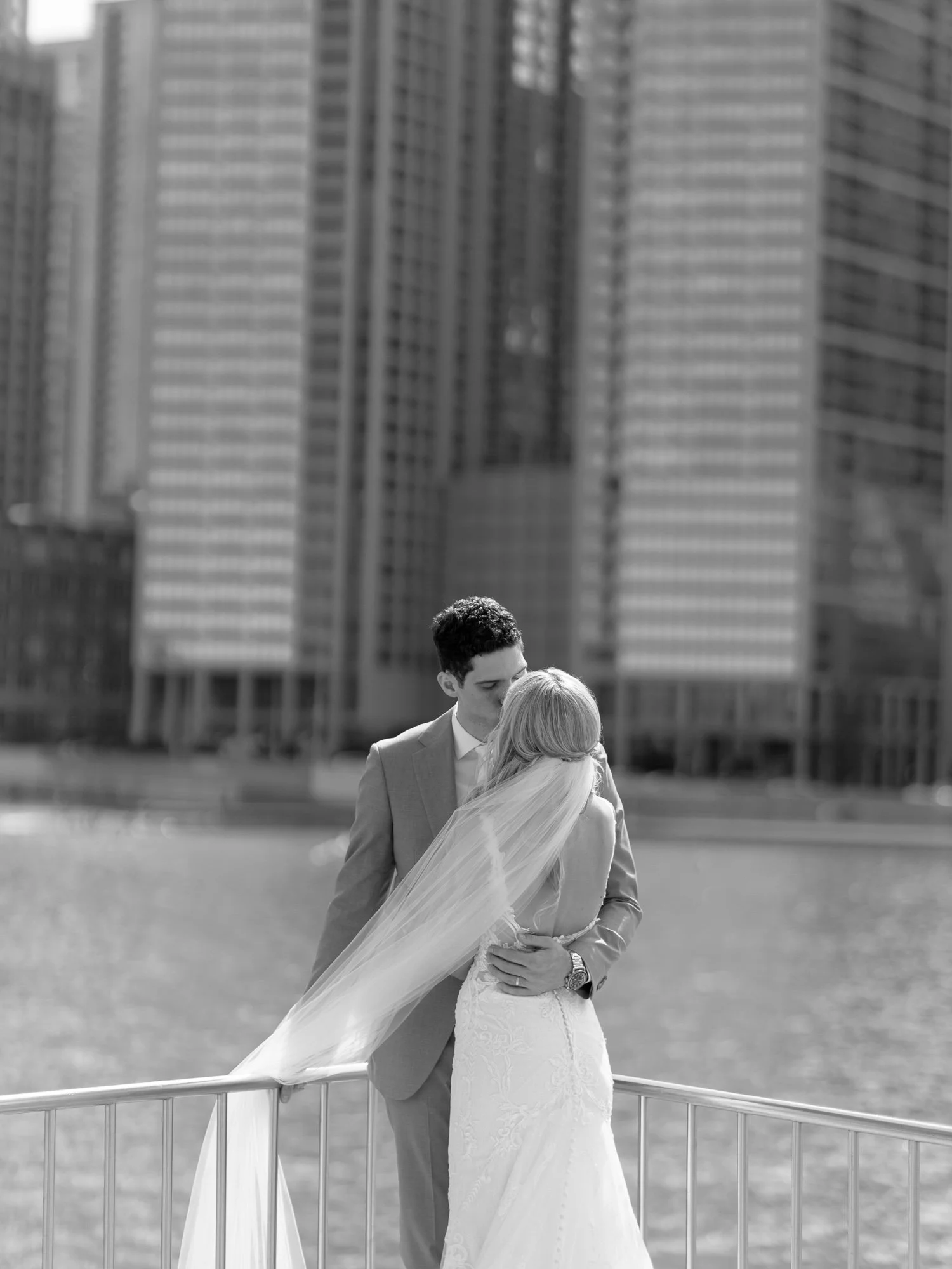 A black-and-white portrait of the bride and groom locked in an intimate embrace along the Chicago Riverwalk, skyscrapers rising dramatically behind them.