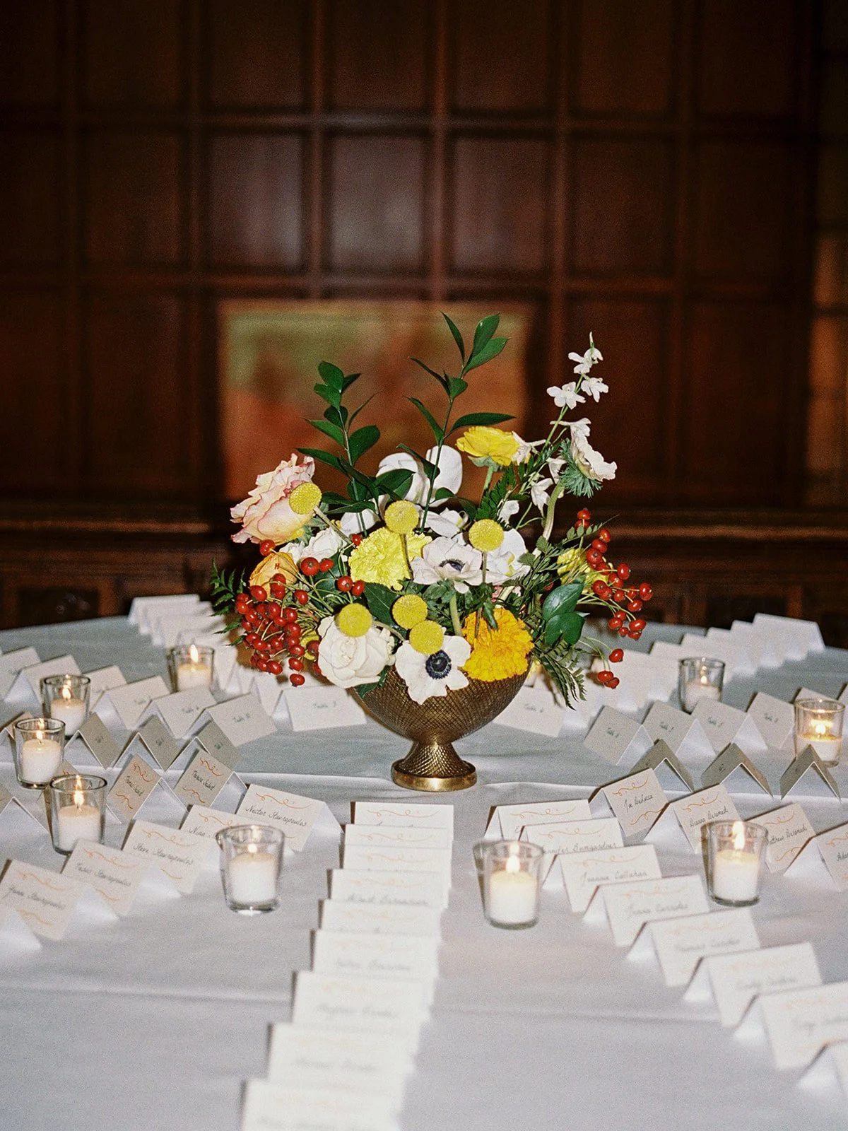 The escort card display at University Club Chicago: a lush floral arrangement of yellow, white, and orange blooms in a brass compote, surrounded by neatly arranged handwritten cards and votives glowing on white linen.