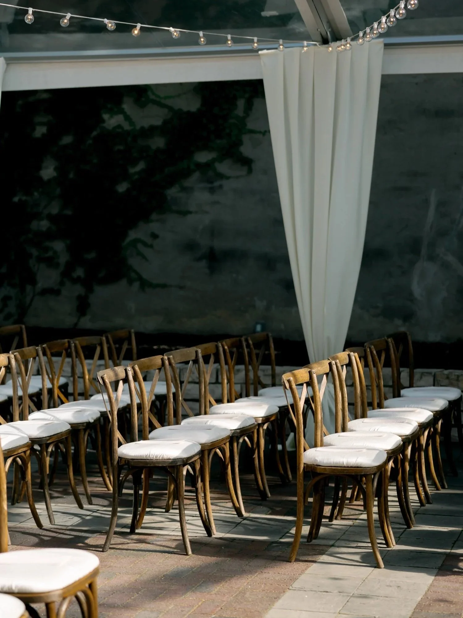 Rows of wooden cross-back chairs await guests under the glass-paneled tent at the Chicago Illuminating Company, white draping and string lights overhead.