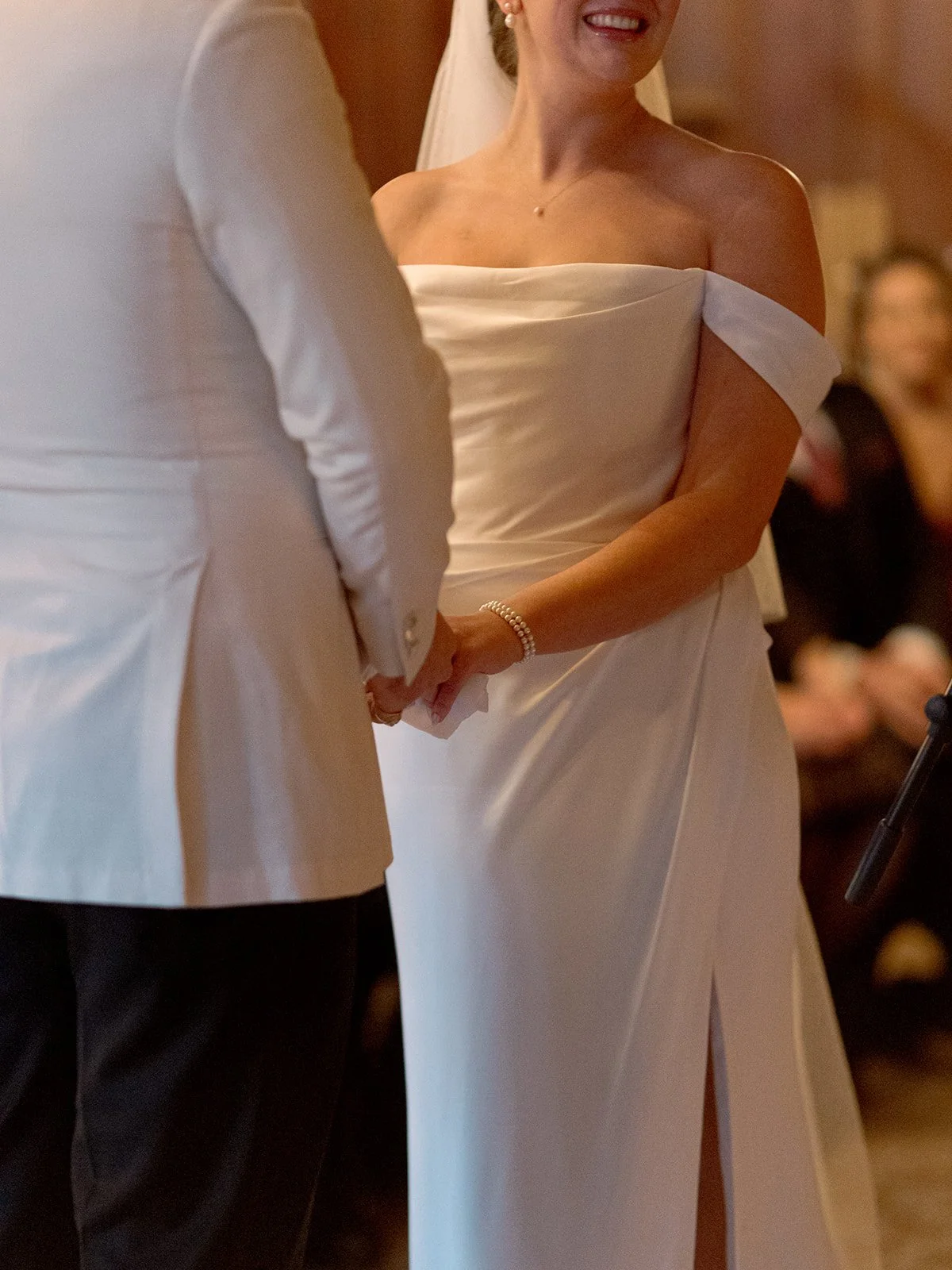 A close-up from the ceremony: the bride's hands clasped with the groom's, a pearl bracelet at her wrist, a wide, uncontainable smile spreading across her face.