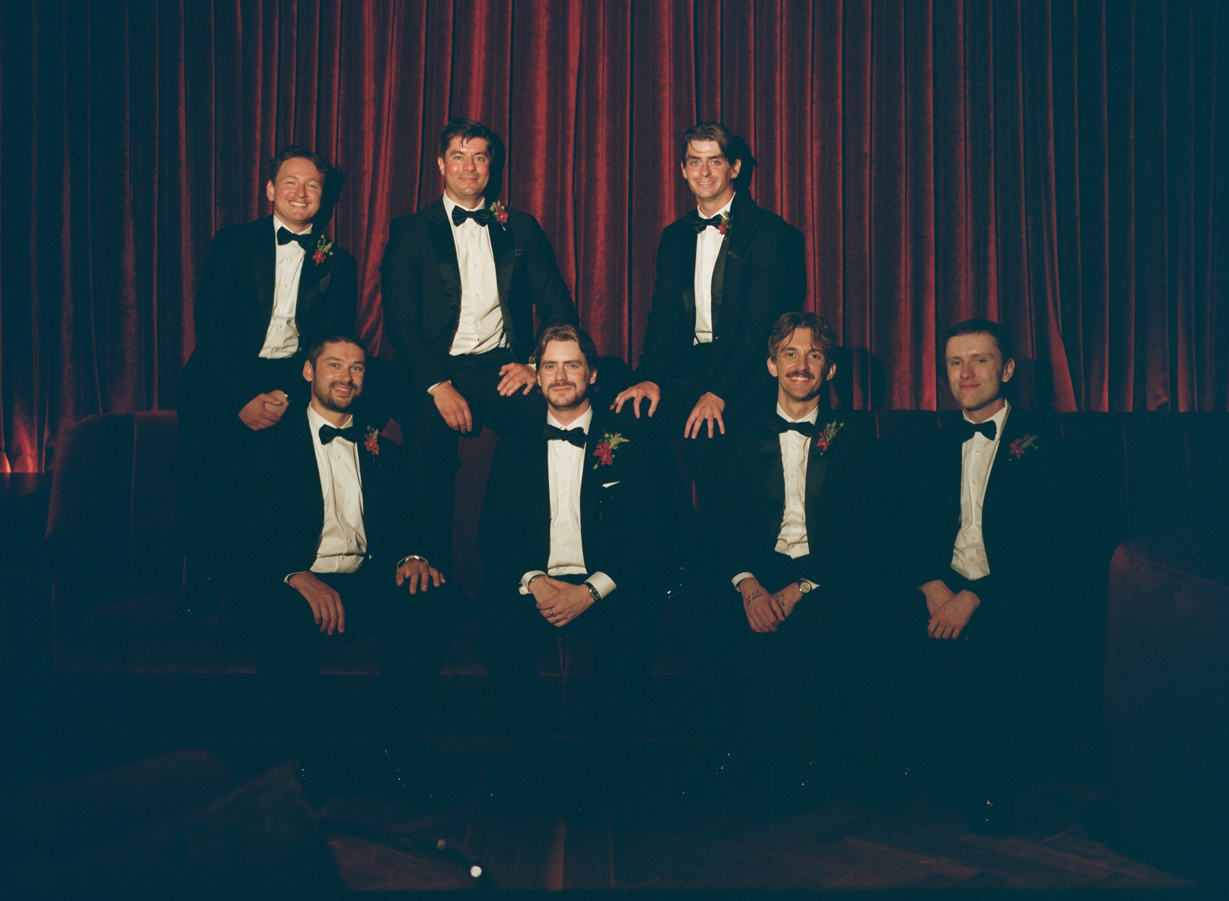 Groomsmen pose together in tuxedos inside The Wellsley's red velvet lounge