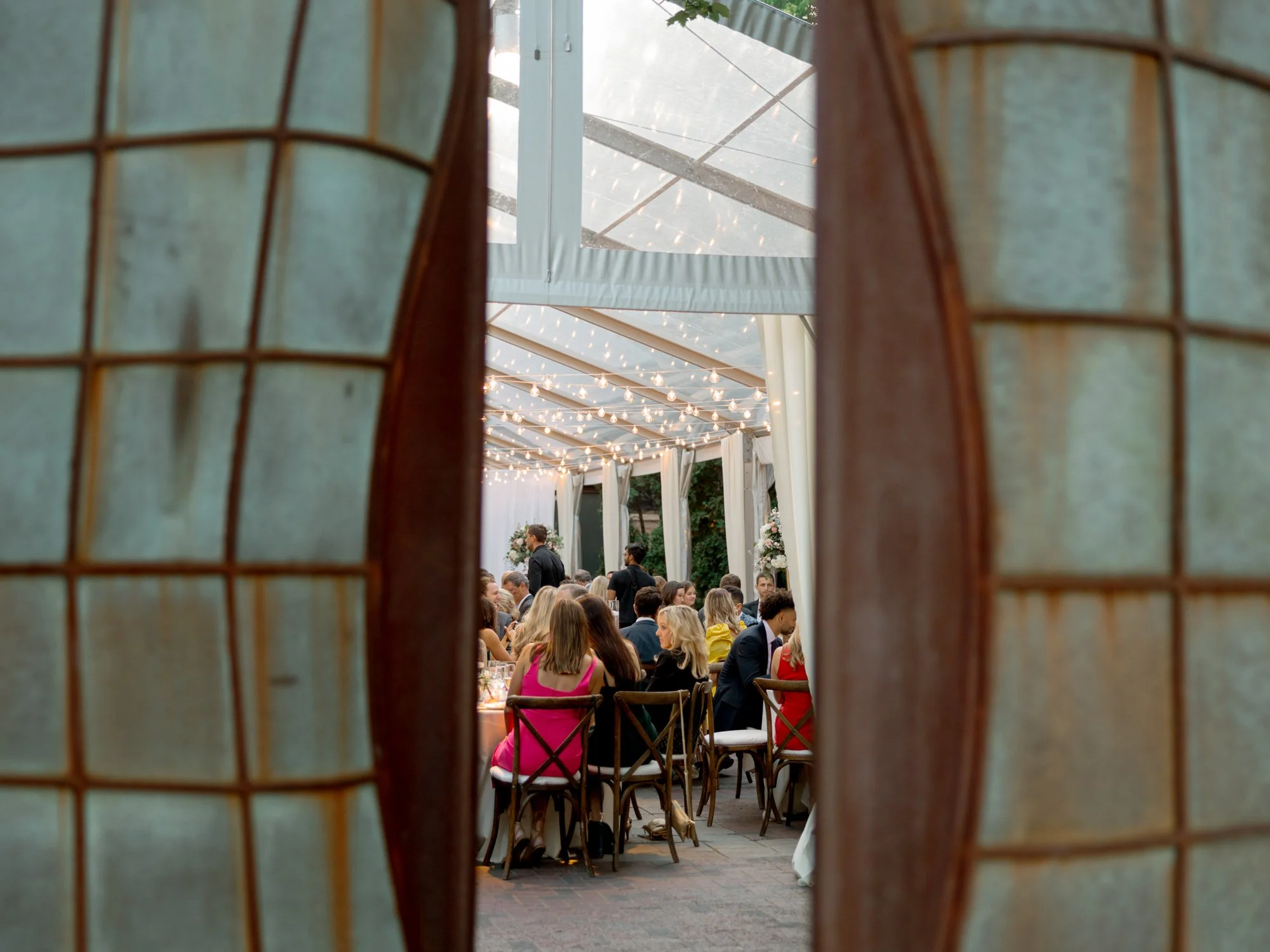 The Chicago Illuminating Company reception glimpsed through ornate iron doors, guests gathered under warm string lights in the garden tent at dusk.