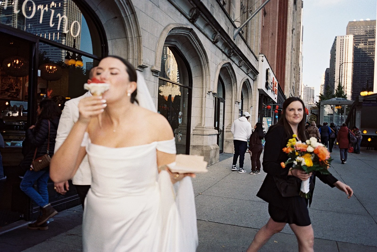 The bride takes a full, unapologetic bite of an ice cream cone on a busy Chicago sidewalk — veil blowing, bridesmaid scrambling behind her with the bouquet.