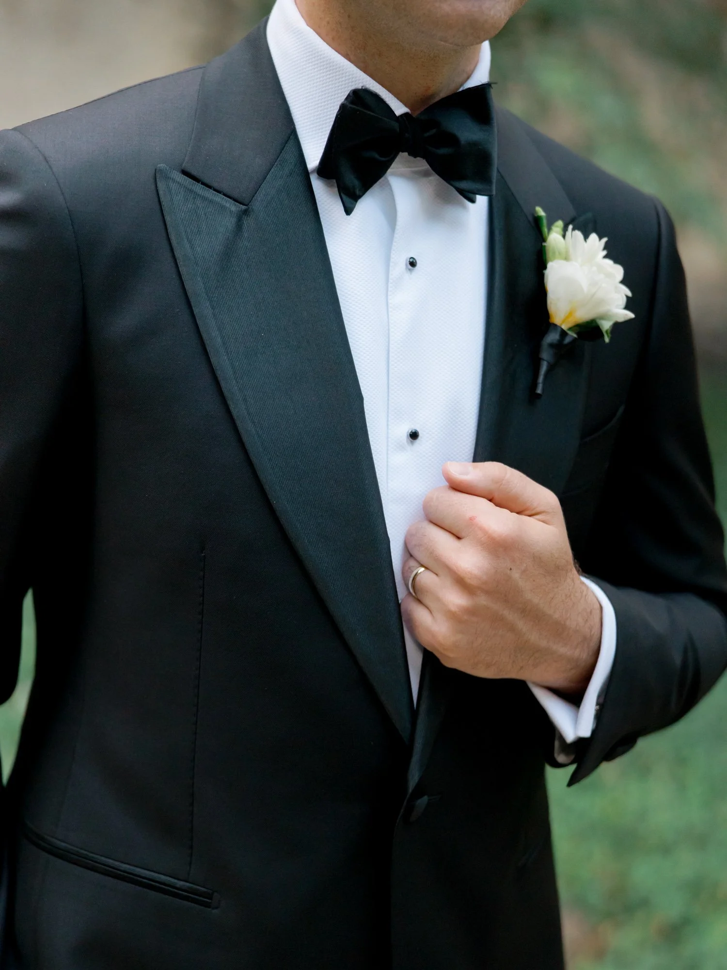 Close-up of the groom’s peak-lapel tuxedo, crisp white boutonniere, and wedding ring hand resting on his chest — the kind of detail that says everything.
