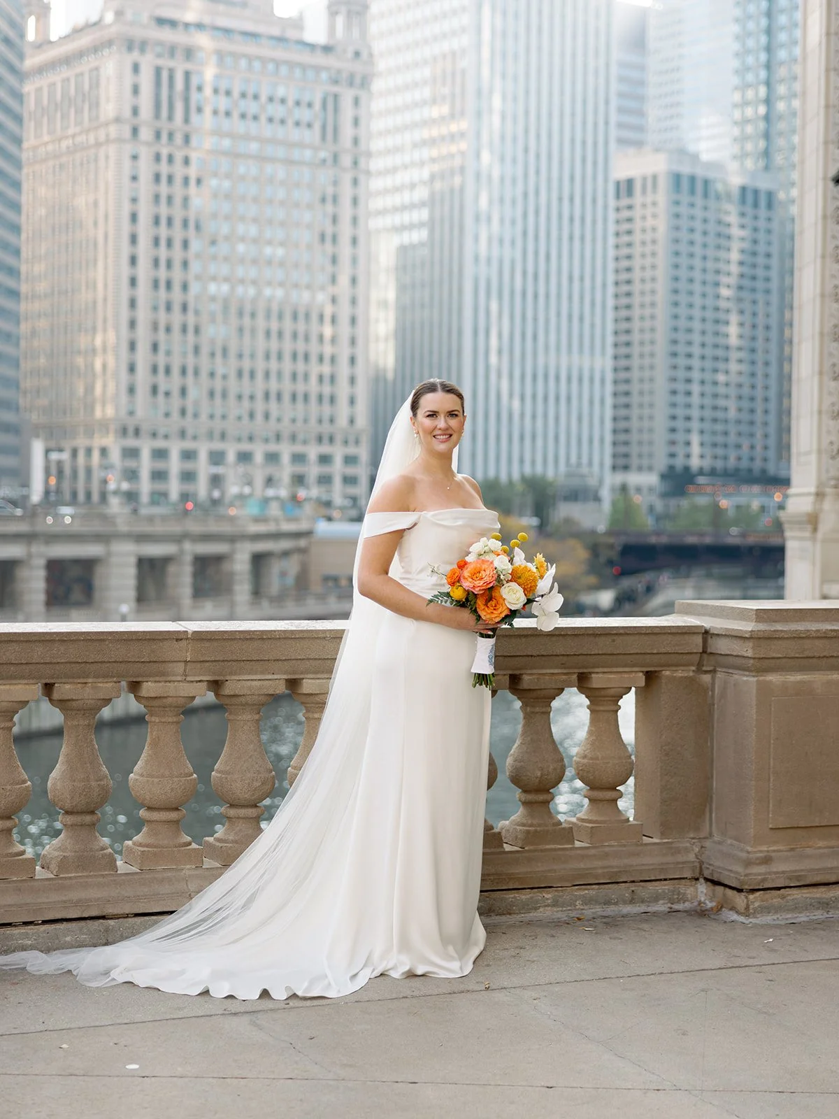 A full-length bridal portrait on the Chicago Riverwalk terrace — cathedral veil, off-shoulder column gown, colorful bouquet, and the entire downtown skyline stretching behind her.