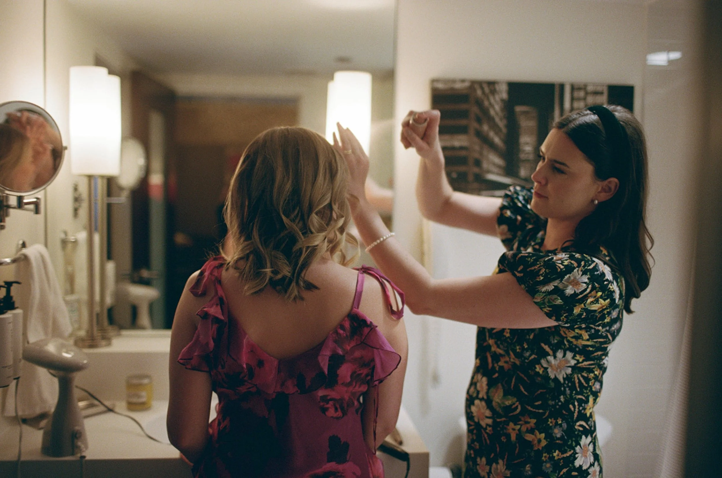 In the bathroom mirror, one bridesmaid carefully fixes the other's hair in the warm glow of hotel lights — a getting-ready candid full of quiet care and anticipation