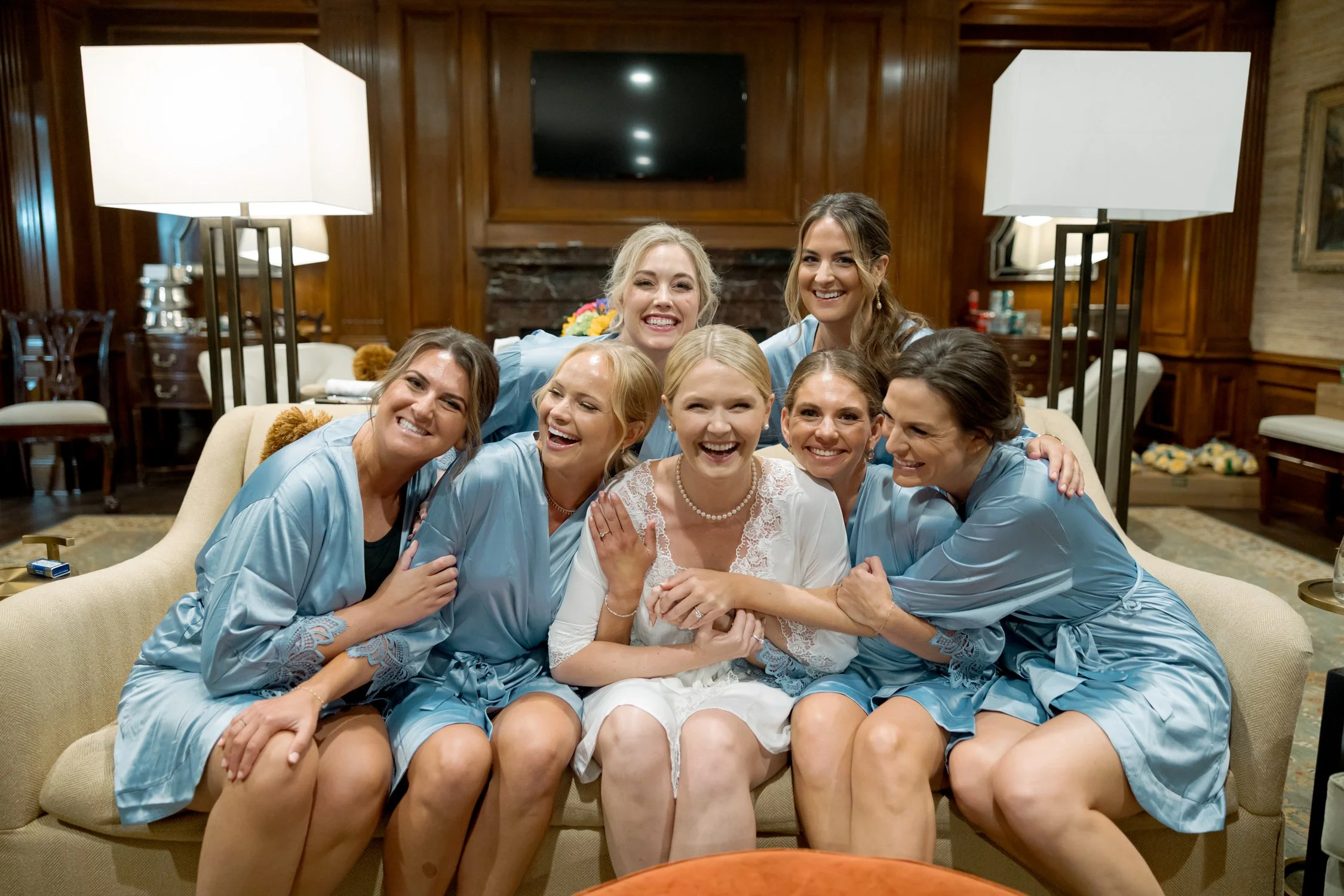 The bride and six bridesmaids crowd onto a sofa in matching light blue robes, laughing together in a portrait that looks exactly like a real memory.
