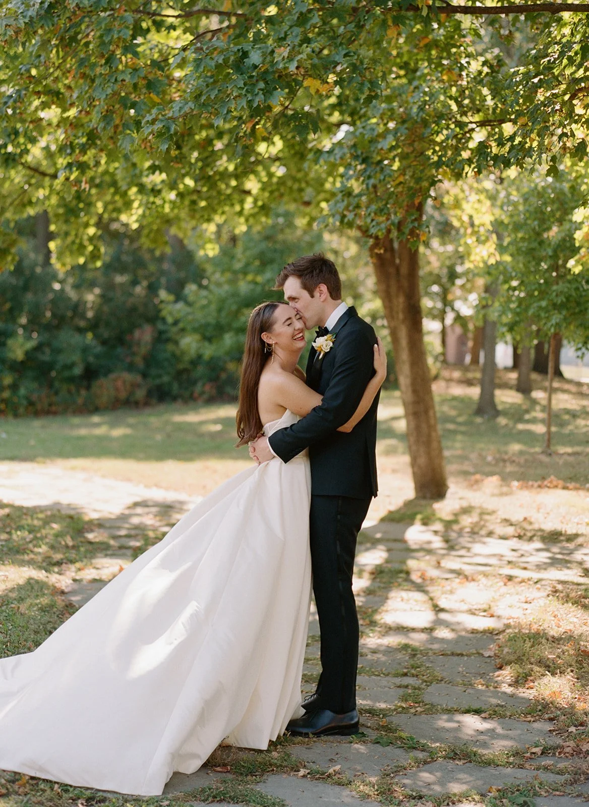 A tender, laughing moment on a stone garden pathway — the groom nuzzling his bride's cheek as her ballgown train sweeps behind them through the lush grounds of Columbus Park Refectory. Chicago film wedding photographer.