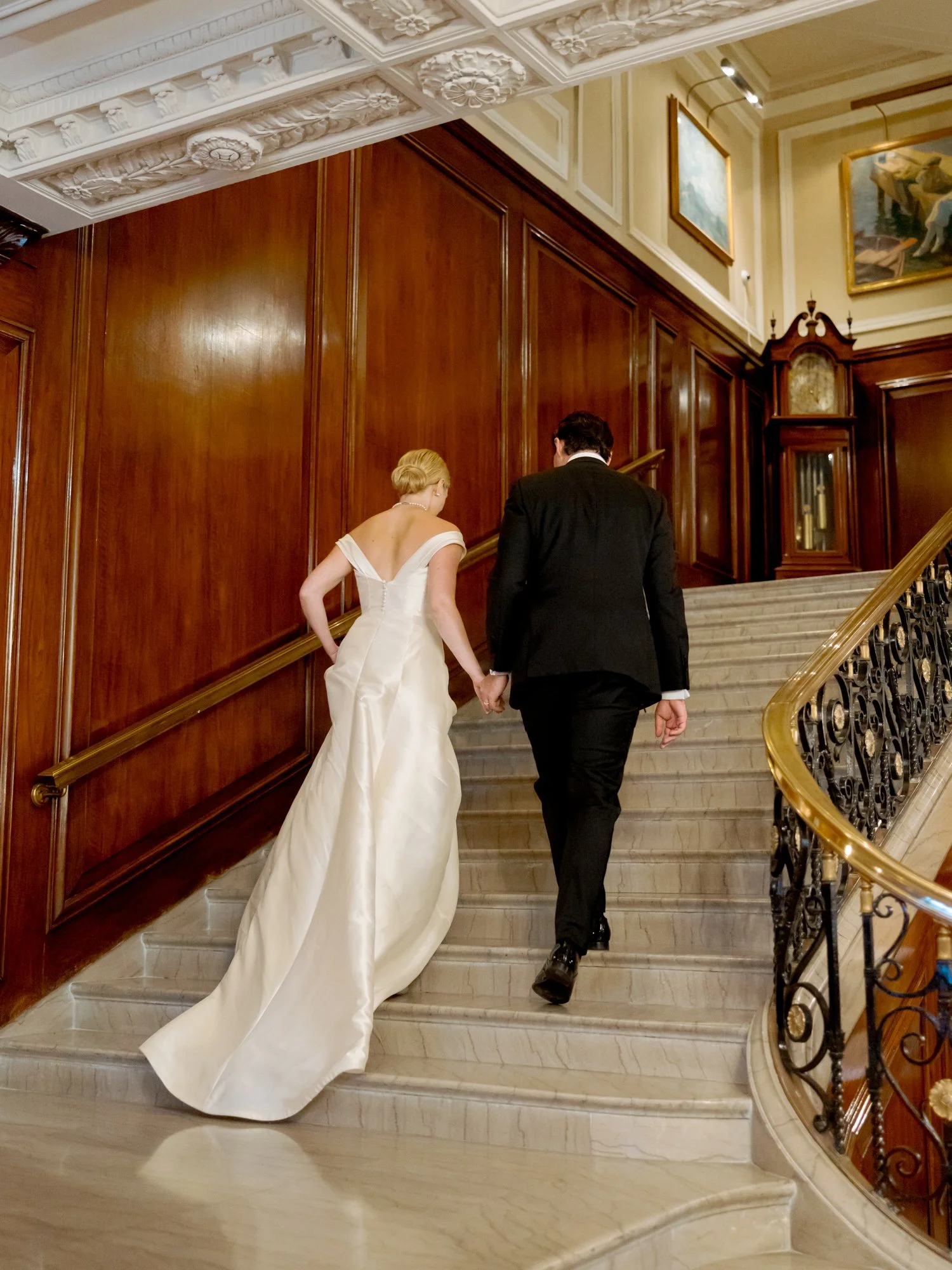 Bride and groom climb the ornate Union League Club grand staircase, the groom reaching back to gather her silk train — dark wood and oil paintings around them.
