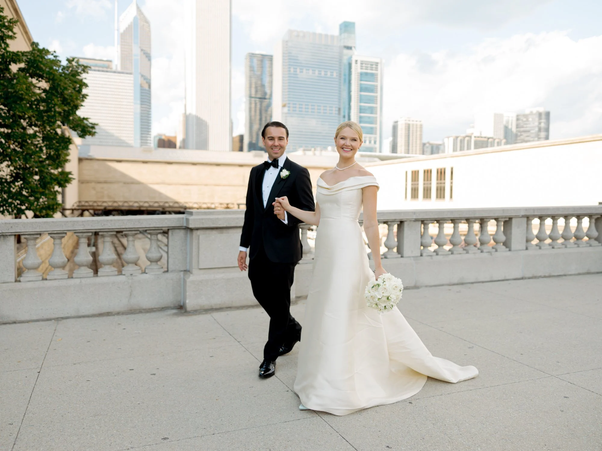 Bride and groom hold hands on the neoclassical stone terrace with the full Chicago skyline rising behind them — clean, classic, unmistakably Chicago.