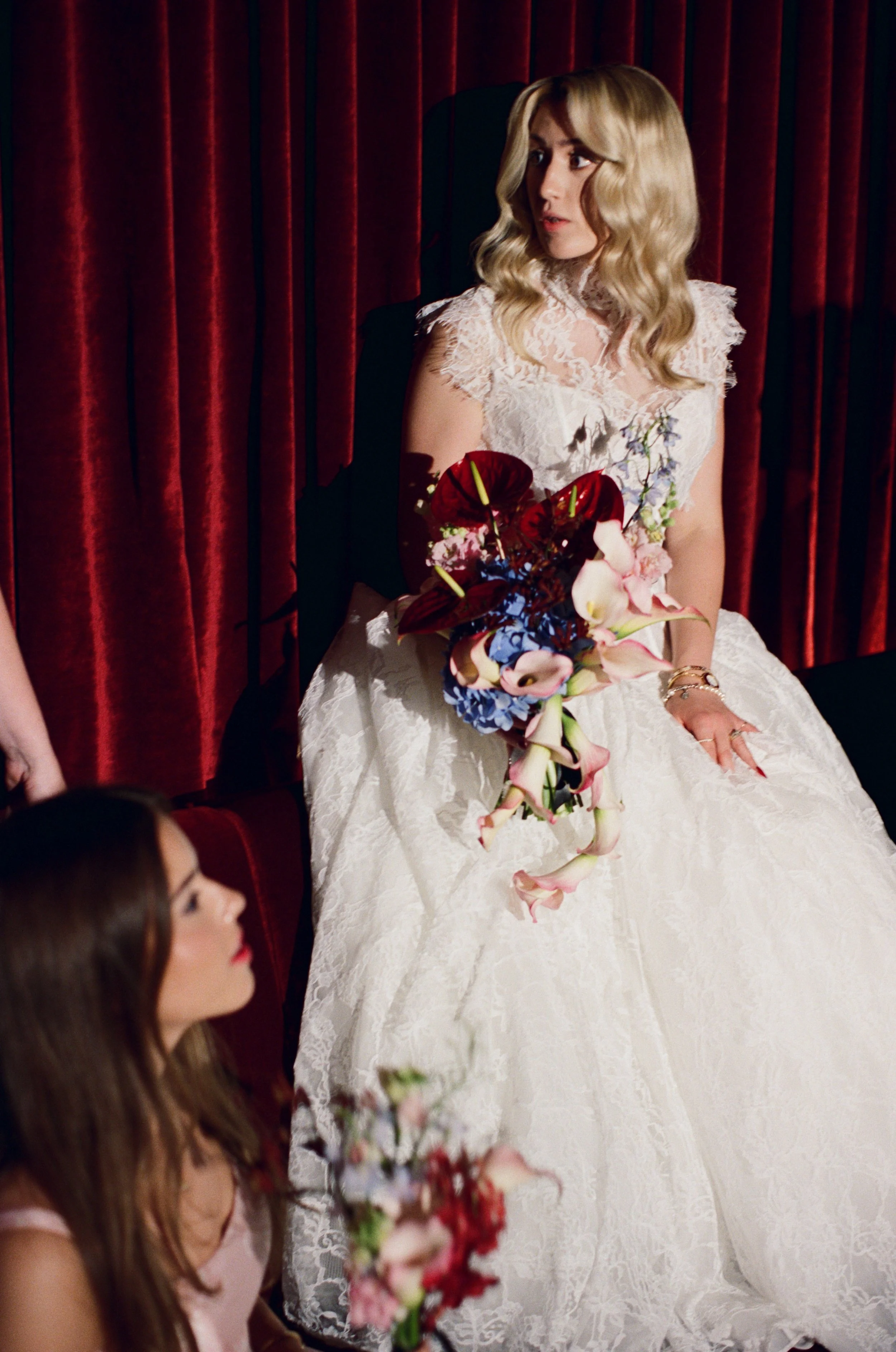 Bride poses alone on the stage inside The Wellsley's red velvet lounge holding a dramatic red and blue bouquet