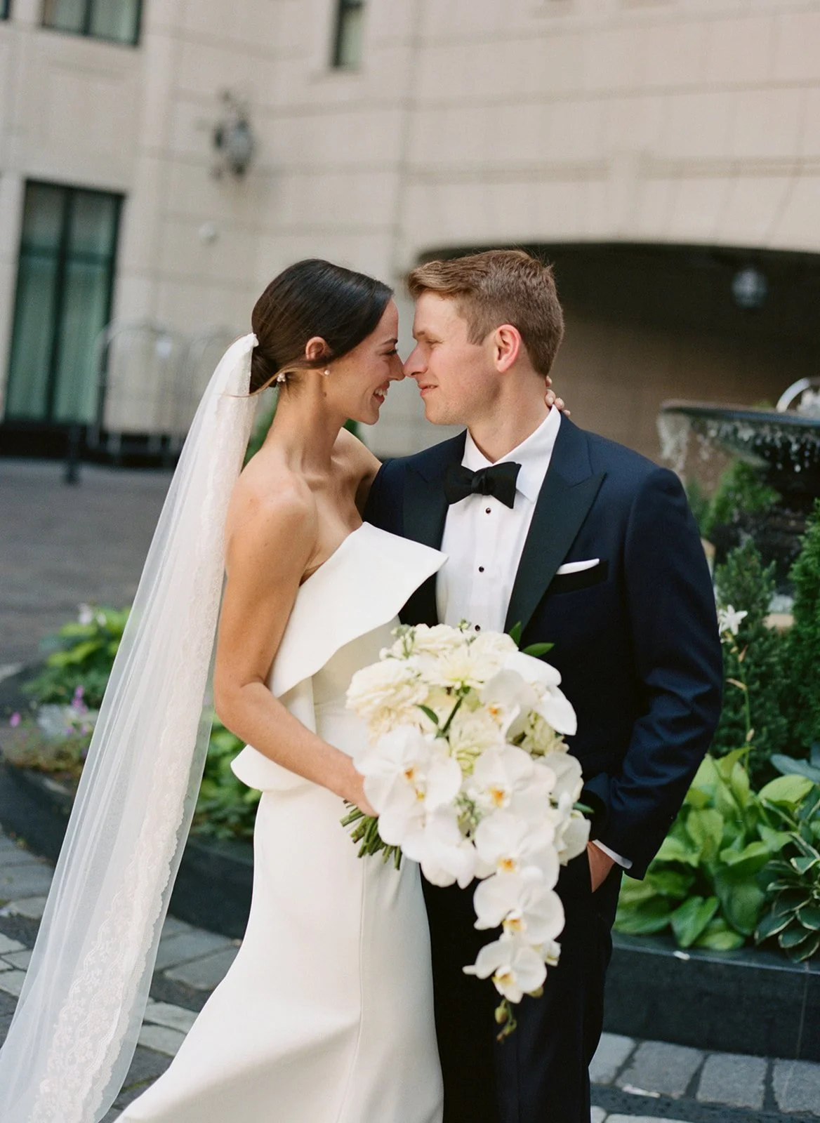 Bride in a structured strapless gown and cathedral veil nearly touching foreheads with her groom in a navy tuxedo in the courtyard of the Waldorf Astoria Chicago, photographed by Louie Abellera.