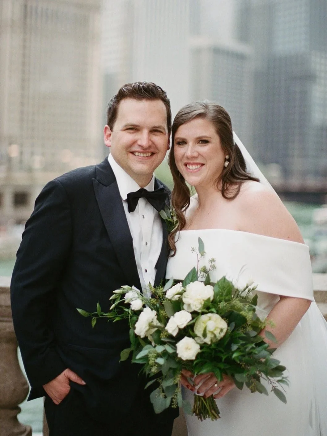 Close portrait of the bride and groom smiling together on the Chicago Riverwalk, downtown skyline in soft focus behind.